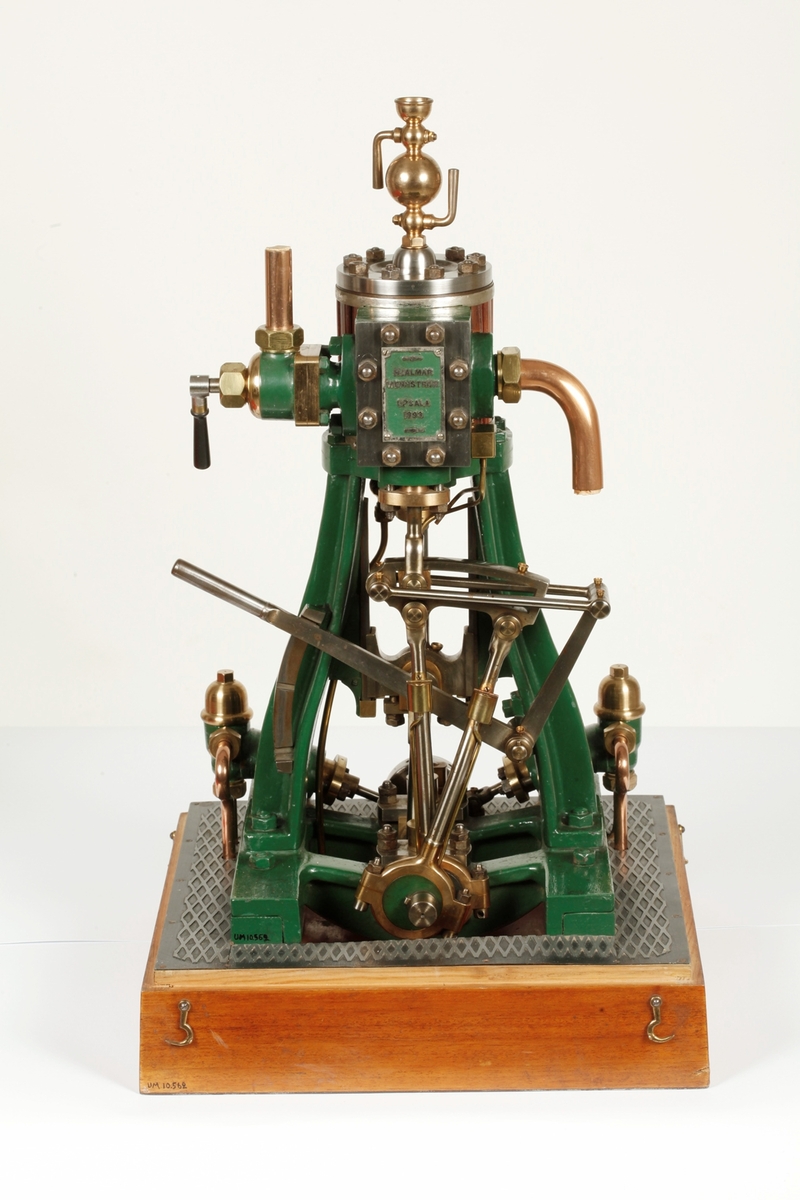 Ångmaskin med grönt järnstativ och övriga delar av stål, mässing och koppar. Skylt på maskinen med text HJALMAR WENNSTRÖM UPSALA 1893. Den står på en fyrkantig bas av polerat trä och är täckt med en rektangulär huv av glas och träribbor fäst med mässingskrokar på basen. Till ångmaskinen hör en "nyckel" (b).