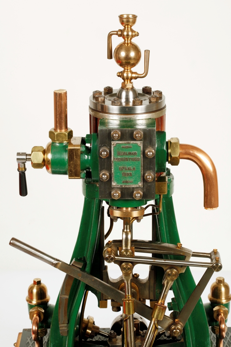 Ångmaskin med grönt järnstativ och övriga delar av stål, mässing och koppar. Skylt på maskinen med text HJALMAR WENNSTRÖM UPSALA 1893. Den står på en fyrkantig bas av polerat trä och är täckt med en rektangulär huv av glas och träribbor fäst med mässingskrokar på basen. Till ångmaskinen hör en "nyckel" (b).