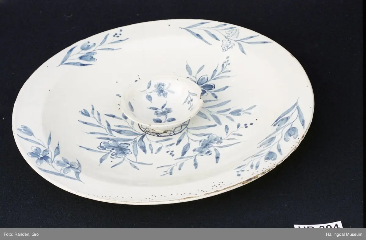 Fajanse, melkehvitt, rundt. Undersiden vid, markert fotring. Svakt skrånende side, smal rand. Dekorasjoner i blått, omkring smørgruben 3 grener med smale blad og hver en blomst. Langs randen 4 grener med forskjellige blomster. R.B.S.
