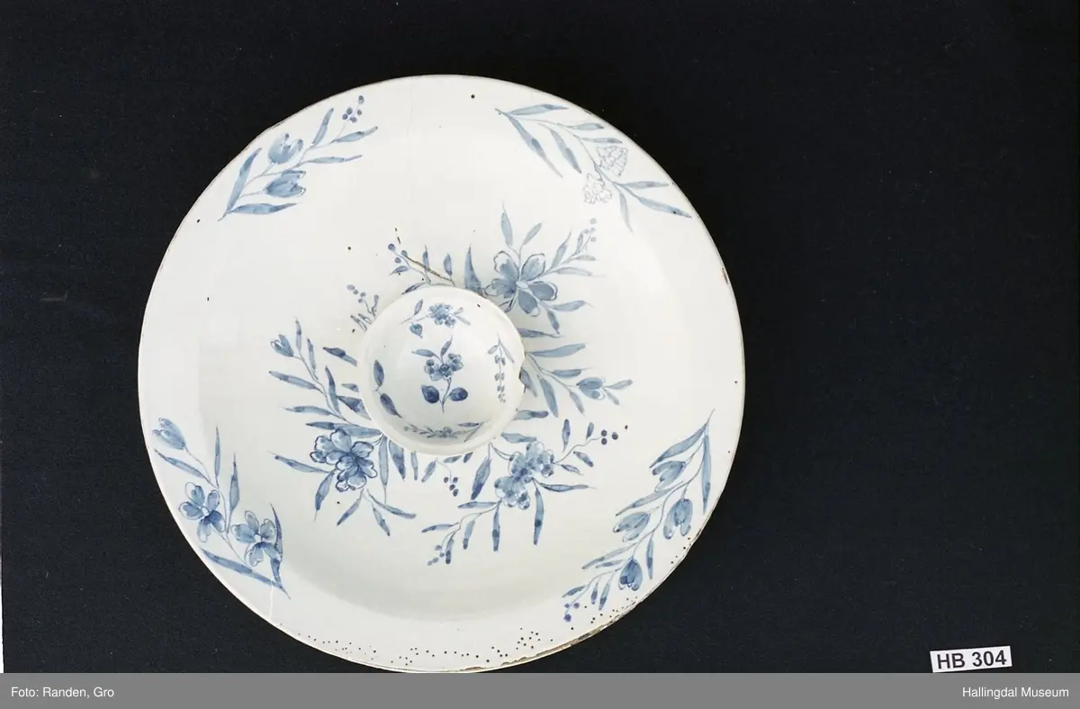 Fajanse, melkehvitt, rundt. Undersiden vid, markert fotring. Svakt skrånende side, smal rand. Dekorasjoner i blått, omkring smørgruben 3 grener med smale blad og hver en blomst. Langs randen 4 grener med forskjellige blomster. R.B.S.