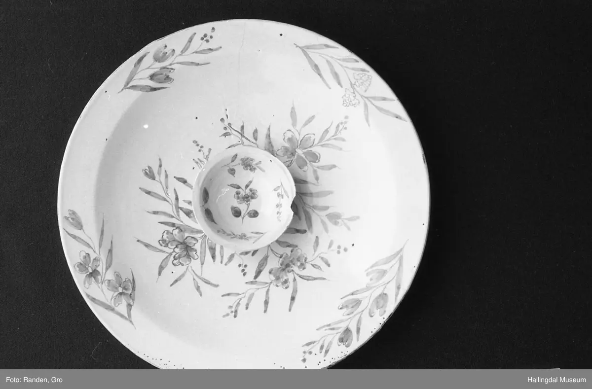 Fajanse, melkehvitt, rundt. Undersiden vid, markert fotring. Svakt skrånende side, smal rand. Dekorasjoner i blått, omkring smørgruben 3 grener med smale blad og hver en blomst. Langs randen 4 grener med forskjellige blomster. R.B.S.