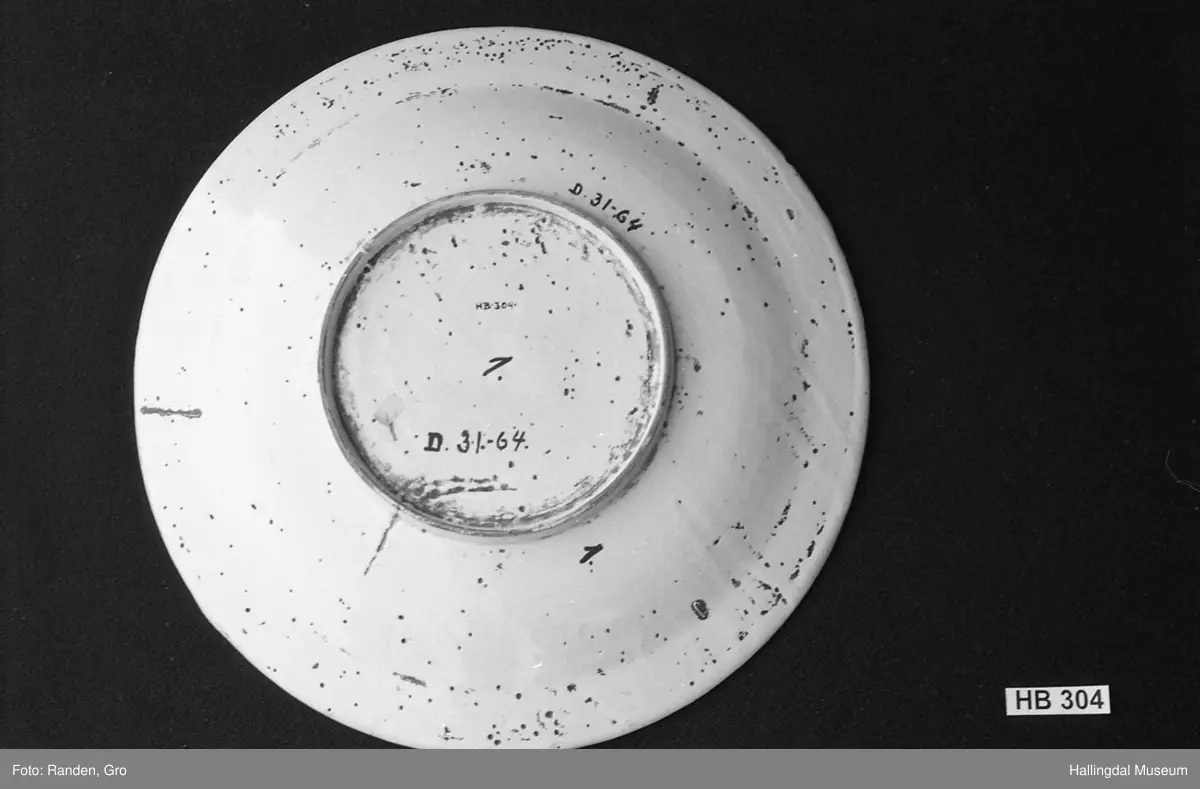 Fajanse, melkehvitt, rundt. Undersiden vid, markert fotring. Svakt skrånende side, smal rand. Dekorasjoner i blått, omkring smørgruben 3 grener med smale blad og hver en blomst. Langs randen 4 grener med forskjellige blomster. R.B.S.