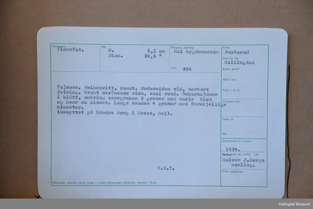 Fajanse, melkehvitt, rundt. Undersiden vid, markert fotring. Svakt skrånende side, smal rand. Dekorasjoner i blått, omkring smørgruben 3 grener med smale blad og hver en blomst. Langs randen 4 grener med forskjellige blomster. R.B.S.