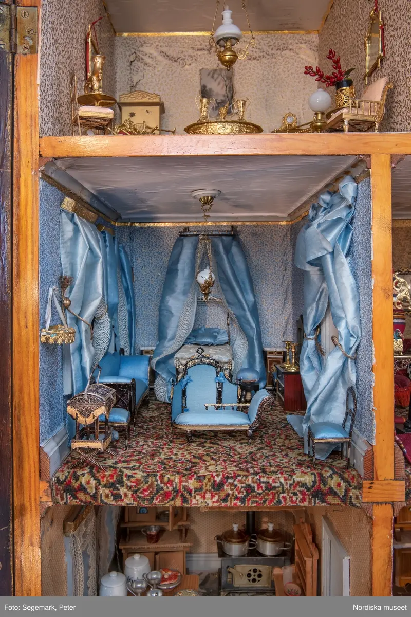 Inventering Sesam 1996-1999:
L 79 cm, B 46 cm, H 174 cm
Dockskåp av brunbetsat furu. Skåp med ett par glasdörrar med brun dekorrand på ramarna, undertill en låda med två dragknoppar, rakt krön med profilerad list. Skåpet står på fyra kulfötter. Dörrparet med lås och nyckel. På gavlarna 4 st fönster på vardera sidan. I skåpet 4 st våningar med sammanlagt 7 rum. Samtliga rum med mönstrade papperstapeter och krok i taket för lampa.
VÅNING 1: 1 rum, heltäckningsmatta av blåmönstrat tyg på vit botten. På höger kortvägg 3 st små pappersbilder i färgtryckav liten flicka i olika situationer. På  bakväggen en europakarta med tysk text.
VÅNING 2:  2 st rum. Bägge med mönstrade linoleummattor och två fönster. Gråmålad dörr mellan rummen. Vänster rum; Spiskåpa av vit papp på bakväggen. 2 st gardinlufter av vit spets med kornischer av gulmetall med påklistrad guldpappersremsa med mönster i relief. Höger rum; två gardinlufter av brunrandigt tyg, kornischer som ovan, omtag av metallkedjor på ena luften och rosa sidenband på andra. Ovanför dörren sitter 3 st beslag av pressad plåt. En större med måne och blommor och två mindre med sköld och blommor. 
VÅNING 3: bägge rummen med mönstrad heltäckningsmatta med lugg och två fönster. Vänster rum; 2 gardinlufter, 1 sänghimmel och 1 draperi av blått siden med vit kantspets. Gardiner med kornischer som ovan. 5 st omtag i form av snodd av gulmetall. Höger rum; enfärgade röda tapeter,2 st gardinlufter av rödrandigt siden med omtag av samma tyg, undergardin av vit spets, kornischer som ovan. Ett draperi av rött och randigt (i flera färger) ylle med ett omtag av metallkedja.
VÅNING 4: Vänster rum; flerfärgat och småmönstrad linoleummatta, Höger rum; utan golvbeläggning. 
- Ett par gröna gardiner av stärkt bomull, som ursprunglingen har suttit innanför glasdörrarna på dockskåpet. Rynkade i båda kortändorna. B 46,5, L 128 cm
Har tillhört givaren. (född 1886 enl. adelskalendern). 
Inventarier till dockskåpet inv.nr 164571-164728
Leif Wallin nov 1997