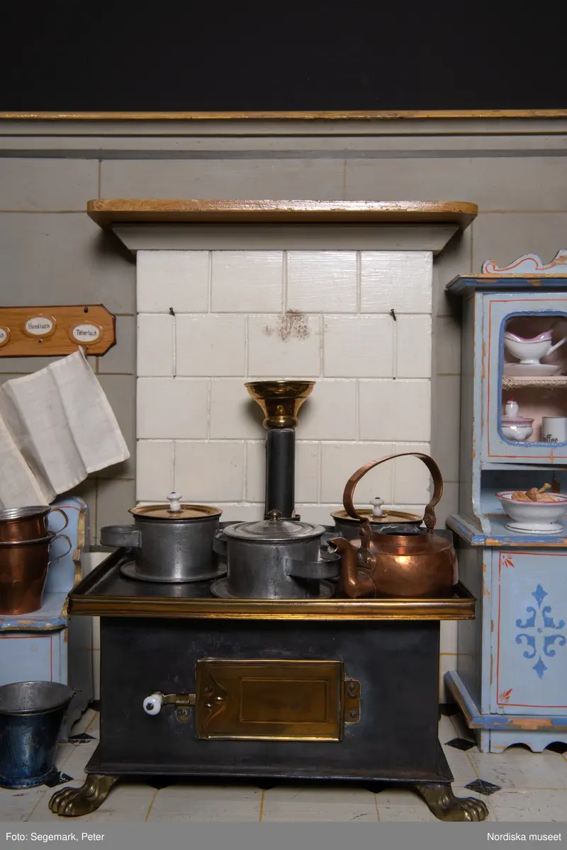 Inventering Sesam 1996-1999:
L 88  H 43,5  Dj 44   cm
Dockskåp, köksdockskåp, av trä, skafferi och kök, utvändigt träådrat. Väggarna i köket kvadermålade och golvet svart och vitrutigt. Skafferiets väggar rosa, väggfasta hyllor, dörrposten ekimiterad.
Lös inredning se inv. 192.992-193.058
Birgitta Martinius 1996