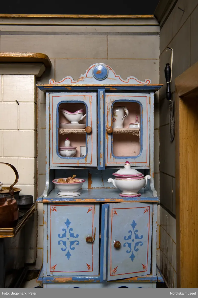 Inventering Sesam 1996-1999:
L 88  H 43,5  Dj 44   cm
Dockskåp, köksdockskåp, av trä, skafferi och kök, utvändigt träådrat. Väggarna i köket kvadermålade och golvet svart och vitrutigt. Skafferiets väggar rosa, väggfasta hyllor, dörrposten ekimiterad.
Lös inredning se inv. 192.992-193.058
Birgitta Martinius 1996