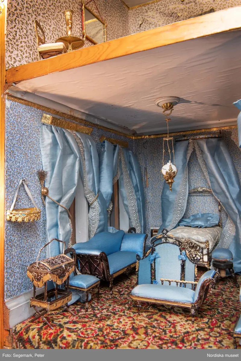 Inventering Sesam 1996-1999:
L 79 cm, B 46 cm, H 174 cm
Dockskåp av brunbetsat furu. Skåp med ett par glasdörrar med brun dekorrand på ramarna, undertill en låda med två dragknoppar, rakt krön med profilerad list. Skåpet står på fyra kulfötter. Dörrparet med lås och nyckel. På gavlarna 4 st fönster på vardera sidan. I skåpet 4 st våningar med sammanlagt 7 rum. Samtliga rum med mönstrade papperstapeter och krok i taket för lampa.
VÅNING 1: 1 rum, heltäckningsmatta av blåmönstrat tyg på vit botten. På höger kortvägg 3 st små pappersbilder i färgtryckav liten flicka i olika situationer. På  bakväggen en europakarta med tysk text.
VÅNING 2:  2 st rum. Bägge med mönstrade linoleummattor och två fönster. Gråmålad dörr mellan rummen. Vänster rum; Spiskåpa av vit papp på bakväggen. 2 st gardinlufter av vit spets med kornischer av gulmetall med påklistrad guldpappersremsa med mönster i relief. Höger rum; två gardinlufter av brunrandigt tyg, kornischer som ovan, omtag av metallkedjor på ena luften och rosa sidenband på andra. Ovanför dörren sitter 3 st beslag av pressad plåt. En större med måne och blommor och två mindre med sköld och blommor. 
VÅNING 3: bägge rummen med mönstrad heltäckningsmatta med lugg och två fönster. Vänster rum; 2 gardinlufter, 1 sänghimmel och 1 draperi av blått siden med vit kantspets. Gardiner med kornischer som ovan. 5 st omtag i form av snodd av gulmetall. Höger rum; enfärgade röda tapeter,2 st gardinlufter av rödrandigt siden med omtag av samma tyg, undergardin av vit spets, kornischer som ovan. Ett draperi av rött och randigt (i flera färger) ylle med ett omtag av metallkedja.
VÅNING 4: Vänster rum; flerfärgat och småmönstrad linoleummatta, Höger rum; utan golvbeläggning. 
- Ett par gröna gardiner av stärkt bomull, som ursprunglingen har suttit innanför glasdörrarna på dockskåpet. Rynkade i båda kortändorna. B 46,5, L 128 cm
Har tillhört givaren. (född 1886 enl. adelskalendern). 
Inventarier till dockskåpet inv.nr 164571-164728
Leif Wallin nov 1997