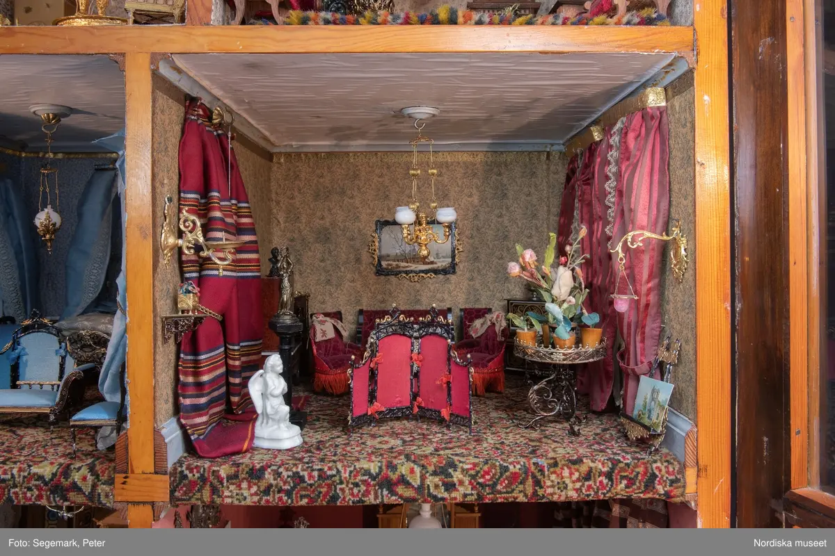 Inventering Sesam 1996-1999:
L 79 cm, B 46 cm, H 174 cm
Dockskåp av brunbetsat furu. Skåp med ett par glasdörrar med brun dekorrand på ramarna, undertill en låda med två dragknoppar, rakt krön med profilerad list. Skåpet står på fyra kulfötter. Dörrparet med lås och nyckel. På gavlarna 4 st fönster på vardera sidan. I skåpet 4 st våningar med sammanlagt 7 rum. Samtliga rum med mönstrade papperstapeter och krok i taket för lampa.
VÅNING 1: 1 rum, heltäckningsmatta av blåmönstrat tyg på vit botten. På höger kortvägg 3 st små pappersbilder i färgtryckav liten flicka i olika situationer. På  bakväggen en europakarta med tysk text.
VÅNING 2:  2 st rum. Bägge med mönstrade linoleummattor och två fönster. Gråmålad dörr mellan rummen. Vänster rum; Spiskåpa av vit papp på bakväggen. 2 st gardinlufter av vit spets med kornischer av gulmetall med påklistrad guldpappersremsa med mönster i relief. Höger rum; två gardinlufter av brunrandigt tyg, kornischer som ovan, omtag av metallkedjor på ena luften och rosa sidenband på andra. Ovanför dörren sitter 3 st beslag av pressad plåt. En större med måne och blommor och två mindre med sköld och blommor. 
VÅNING 3: bägge rummen med mönstrad heltäckningsmatta med lugg och två fönster. Vänster rum; 2 gardinlufter, 1 sänghimmel och 1 draperi av blått siden med vit kantspets. Gardiner med kornischer som ovan. 5 st omtag i form av snodd av gulmetall. Höger rum; enfärgade röda tapeter,2 st gardinlufter av rödrandigt siden med omtag av samma tyg, undergardin av vit spets, kornischer som ovan. Ett draperi av rött och randigt (i flera färger) ylle med ett omtag av metallkedja.
VÅNING 4: Vänster rum; flerfärgat och småmönstrad linoleummatta, Höger rum; utan golvbeläggning. 
- Ett par gröna gardiner av stärkt bomull, som ursprunglingen har suttit innanför glasdörrarna på dockskåpet. Rynkade i båda kortändorna. B 46,5, L 128 cm
Har tillhört givaren. (född 1886 enl. adelskalendern). 
Inventarier till dockskåpet inv.nr 164571-164728
Leif Wallin nov 1997