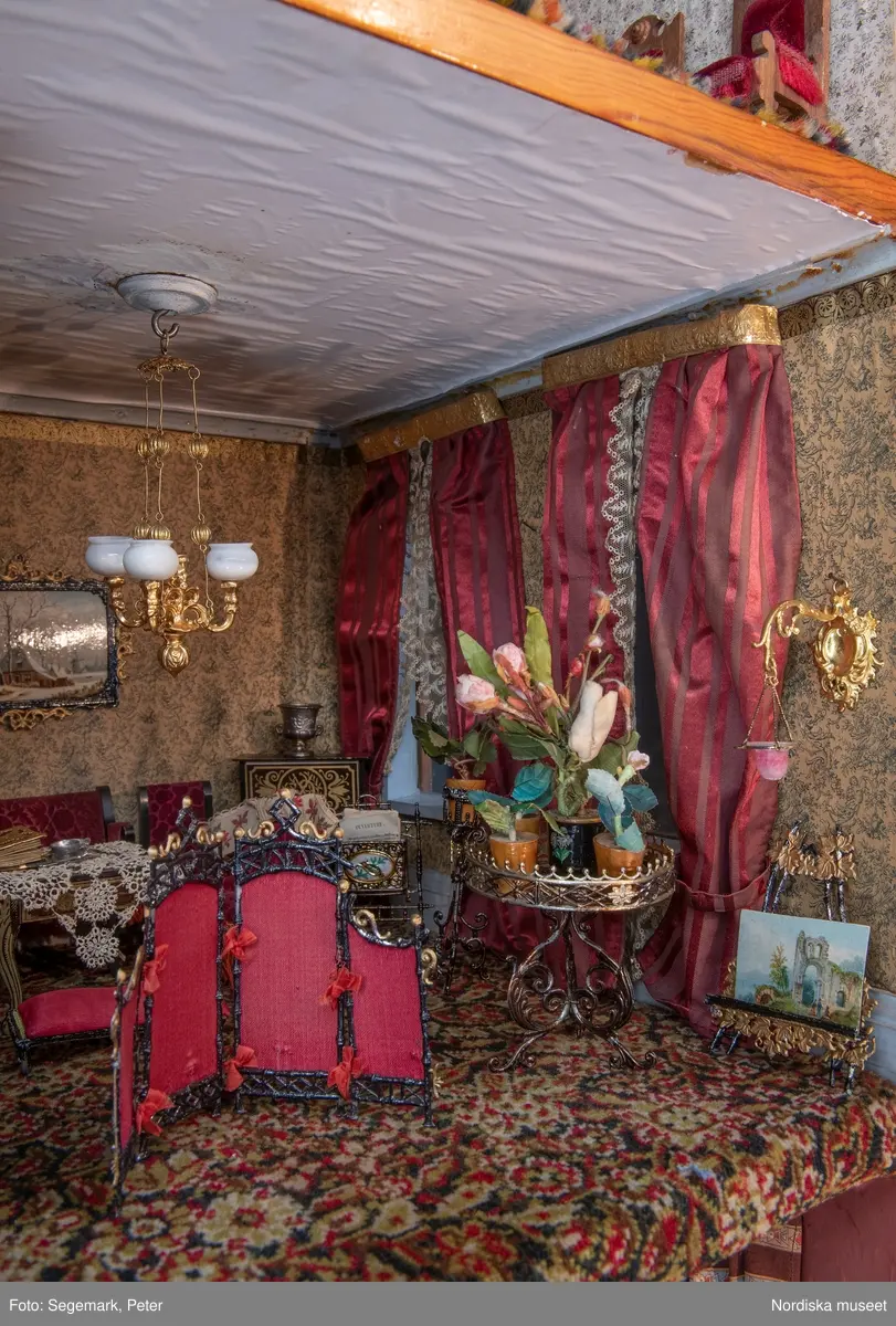 Inventering Sesam 1996-1999:
L 79 cm, B 46 cm, H 174 cm
Dockskåp av brunbetsat furu. Skåp med ett par glasdörrar med brun dekorrand på ramarna, undertill en låda med två dragknoppar, rakt krön med profilerad list. Skåpet står på fyra kulfötter. Dörrparet med lås och nyckel. På gavlarna 4 st fönster på vardera sidan. I skåpet 4 st våningar med sammanlagt 7 rum. Samtliga rum med mönstrade papperstapeter och krok i taket för lampa.
VÅNING 1: 1 rum, heltäckningsmatta av blåmönstrat tyg på vit botten. På höger kortvägg 3 st små pappersbilder i färgtryckav liten flicka i olika situationer. På  bakväggen en europakarta med tysk text.
VÅNING 2:  2 st rum. Bägge med mönstrade linoleummattor och två fönster. Gråmålad dörr mellan rummen. Vänster rum; Spiskåpa av vit papp på bakväggen. 2 st gardinlufter av vit spets med kornischer av gulmetall med påklistrad guldpappersremsa med mönster i relief. Höger rum; två gardinlufter av brunrandigt tyg, kornischer som ovan, omtag av metallkedjor på ena luften och rosa sidenband på andra. Ovanför dörren sitter 3 st beslag av pressad plåt. En större med måne och blommor och två mindre med sköld och blommor. 
VÅNING 3: bägge rummen med mönstrad heltäckningsmatta med lugg och två fönster. Vänster rum; 2 gardinlufter, 1 sänghimmel och 1 draperi av blått siden med vit kantspets. Gardiner med kornischer som ovan. 5 st omtag i form av snodd av gulmetall. Höger rum; enfärgade röda tapeter,2 st gardinlufter av rödrandigt siden med omtag av samma tyg, undergardin av vit spets, kornischer som ovan. Ett draperi av rött och randigt (i flera färger) ylle med ett omtag av metallkedja.
VÅNING 4: Vänster rum; flerfärgat och småmönstrad linoleummatta, Höger rum; utan golvbeläggning. 
- Ett par gröna gardiner av stärkt bomull, som ursprunglingen har suttit innanför glasdörrarna på dockskåpet. Rynkade i båda kortändorna. B 46,5, L 128 cm
Har tillhört givaren. (född 1886 enl. adelskalendern). 
Inventarier till dockskåpet inv.nr 164571-164728
Leif Wallin nov 1997