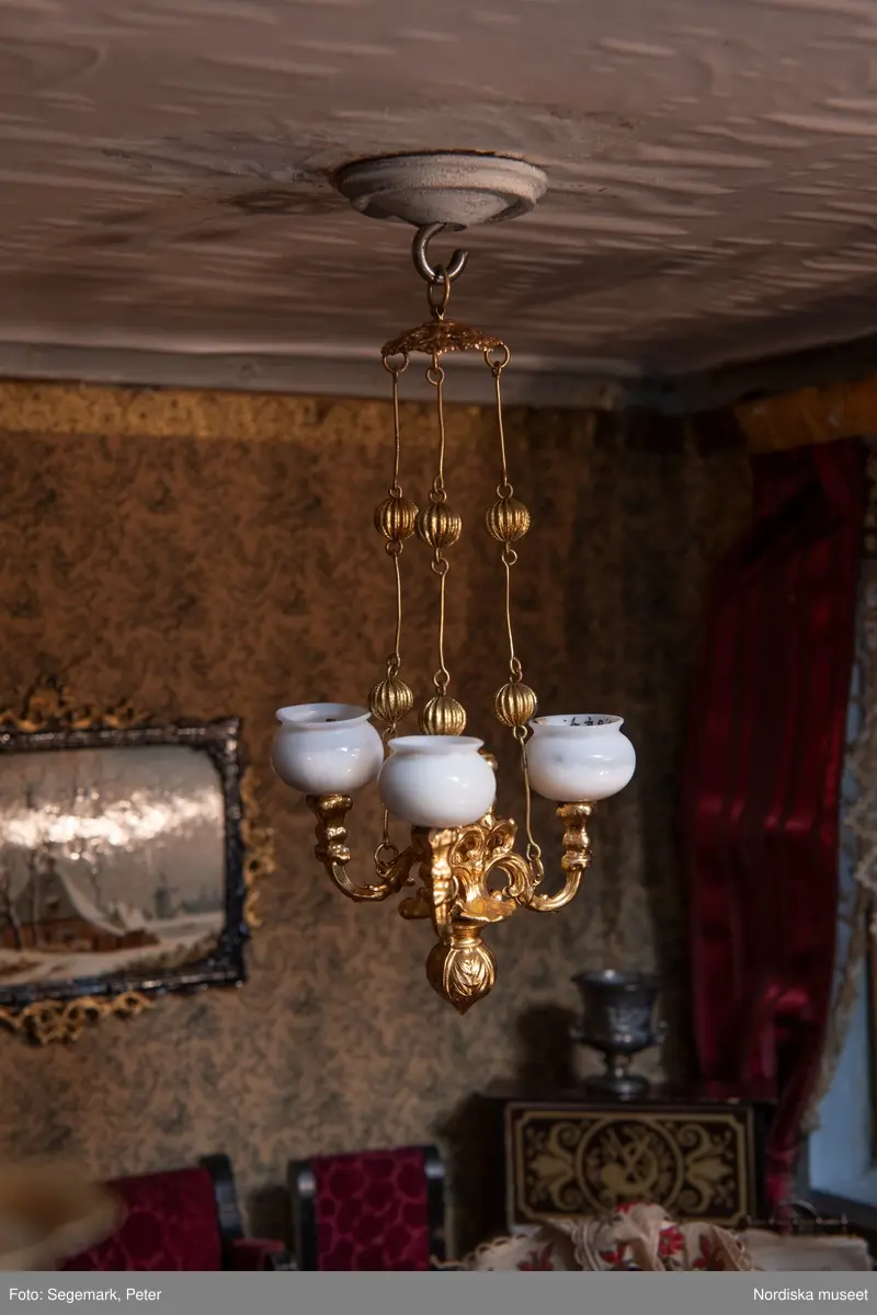 Inventering Sesam 1996-1999:
L 79 cm, B 46 cm, H 174 cm
Dockskåp av brunbetsat furu. Skåp med ett par glasdörrar med brun dekorrand på ramarna, undertill en låda med två dragknoppar, rakt krön med profilerad list. Skåpet står på fyra kulfötter. Dörrparet med lås och nyckel. På gavlarna 4 st fönster på vardera sidan. I skåpet 4 st våningar med sammanlagt 7 rum. Samtliga rum med mönstrade papperstapeter och krok i taket för lampa.
VÅNING 1: 1 rum, heltäckningsmatta av blåmönstrat tyg på vit botten. På höger kortvägg 3 st små pappersbilder i färgtryckav liten flicka i olika situationer. På  bakväggen en europakarta med tysk text.
VÅNING 2:  2 st rum. Bägge med mönstrade linoleummattor och två fönster. Gråmålad dörr mellan rummen. Vänster rum; Spiskåpa av vit papp på bakväggen. 2 st gardinlufter av vit spets med kornischer av gulmetall med påklistrad guldpappersremsa med mönster i relief. Höger rum; två gardinlufter av brunrandigt tyg, kornischer som ovan, omtag av metallkedjor på ena luften och rosa sidenband på andra. Ovanför dörren sitter 3 st beslag av pressad plåt. En större med måne och blommor och två mindre med sköld och blommor. 
VÅNING 3: bägge rummen med mönstrad heltäckningsmatta med lugg och två fönster. Vänster rum; 2 gardinlufter, 1 sänghimmel och 1 draperi av blått siden med vit kantspets. Gardiner med kornischer som ovan. 5 st omtag i form av snodd av gulmetall. Höger rum; enfärgade röda tapeter,2 st gardinlufter av rödrandigt siden med omtag av samma tyg, undergardin av vit spets, kornischer som ovan. Ett draperi av rött och randigt (i flera färger) ylle med ett omtag av metallkedja.
VÅNING 4: Vänster rum; flerfärgat och småmönstrad linoleummatta, Höger rum; utan golvbeläggning. 
- Ett par gröna gardiner av stärkt bomull, som ursprunglingen har suttit innanför glasdörrarna på dockskåpet. Rynkade i båda kortändorna. B 46,5, L 128 cm
Har tillhört givaren. (född 1886 enl. adelskalendern). 
Inventarier till dockskåpet inv.nr 164571-164728
Leif Wallin nov 1997