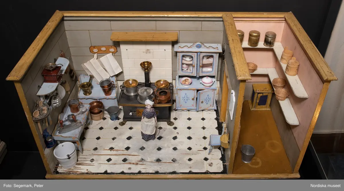 Inventering Sesam 1996-1999:
L 88  H 43,5  Dj 44   cm
Dockskåp, köksdockskåp, av trä, skafferi och kök, utvändigt träådrat. Väggarna i köket kvadermålade och golvet svart och vitrutigt. Skafferiets väggar rosa, väggfasta hyllor, dörrposten ekimiterad.
Lös inredning se inv. 192.992-193.058
Birgitta Martinius 1996