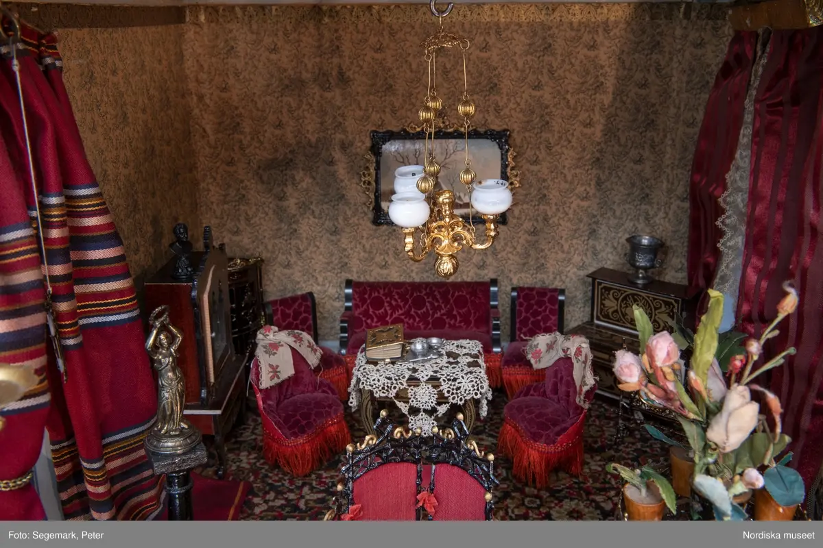 Inventering Sesam 1996-1999:
L 79 cm, B 46 cm, H 174 cm
Dockskåp av brunbetsat furu. Skåp med ett par glasdörrar med brun dekorrand på ramarna, undertill en låda med två dragknoppar, rakt krön med profilerad list. Skåpet står på fyra kulfötter. Dörrparet med lås och nyckel. På gavlarna 4 st fönster på vardera sidan. I skåpet 4 st våningar med sammanlagt 7 rum. Samtliga rum med mönstrade papperstapeter och krok i taket för lampa.
VÅNING 1: 1 rum, heltäckningsmatta av blåmönstrat tyg på vit botten. På höger kortvägg 3 st små pappersbilder i färgtryckav liten flicka i olika situationer. På  bakväggen en europakarta med tysk text.
VÅNING 2:  2 st rum. Bägge med mönstrade linoleummattor och två fönster. Gråmålad dörr mellan rummen. Vänster rum; Spiskåpa av vit papp på bakväggen. 2 st gardinlufter av vit spets med kornischer av gulmetall med påklistrad guldpappersremsa med mönster i relief. Höger rum; två gardinlufter av brunrandigt tyg, kornischer som ovan, omtag av metallkedjor på ena luften och rosa sidenband på andra. Ovanför dörren sitter 3 st beslag av pressad plåt. En större med måne och blommor och två mindre med sköld och blommor. 
VÅNING 3: bägge rummen med mönstrad heltäckningsmatta med lugg och två fönster. Vänster rum; 2 gardinlufter, 1 sänghimmel och 1 draperi av blått siden med vit kantspets. Gardiner med kornischer som ovan. 5 st omtag i form av snodd av gulmetall. Höger rum; enfärgade röda tapeter,2 st gardinlufter av rödrandigt siden med omtag av samma tyg, undergardin av vit spets, kornischer som ovan. Ett draperi av rött och randigt (i flera färger) ylle med ett omtag av metallkedja.
VÅNING 4: Vänster rum; flerfärgat och småmönstrad linoleummatta, Höger rum; utan golvbeläggning. 
- Ett par gröna gardiner av stärkt bomull, som ursprunglingen har suttit innanför glasdörrarna på dockskåpet. Rynkade i båda kortändorna. B 46,5, L 128 cm
Har tillhört givaren. (född 1886 enl. adelskalendern). 
Inventarier till dockskåpet inv.nr 164571-164728
Leif Wallin nov 1997