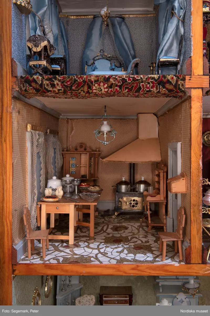 Inventering Sesam 1996-1999:
L 79 cm, B 46 cm, H 174 cm
Dockskåp av brunbetsat furu. Skåp med ett par glasdörrar med brun dekorrand på ramarna, undertill en låda med två dragknoppar, rakt krön med profilerad list. Skåpet står på fyra kulfötter. Dörrparet med lås och nyckel. På gavlarna 4 st fönster på vardera sidan. I skåpet 4 st våningar med sammanlagt 7 rum. Samtliga rum med mönstrade papperstapeter och krok i taket för lampa.
VÅNING 1: 1 rum, heltäckningsmatta av blåmönstrat tyg på vit botten. På höger kortvägg 3 st små pappersbilder i färgtryckav liten flicka i olika situationer. På  bakväggen en europakarta med tysk text.
VÅNING 2:  2 st rum. Bägge med mönstrade linoleummattor och två fönster. Gråmålad dörr mellan rummen. Vänster rum; Spiskåpa av vit papp på bakväggen. 2 st gardinlufter av vit spets med kornischer av gulmetall med påklistrad guldpappersremsa med mönster i relief. Höger rum; två gardinlufter av brunrandigt tyg, kornischer som ovan, omtag av metallkedjor på ena luften och rosa sidenband på andra. Ovanför dörren sitter 3 st beslag av pressad plåt. En större med måne och blommor och två mindre med sköld och blommor. 
VÅNING 3: bägge rummen med mönstrad heltäckningsmatta med lugg och två fönster. Vänster rum; 2 gardinlufter, 1 sänghimmel och 1 draperi av blått siden med vit kantspets. Gardiner med kornischer som ovan. 5 st omtag i form av snodd av gulmetall. Höger rum; enfärgade röda tapeter,2 st gardinlufter av rödrandigt siden med omtag av samma tyg, undergardin av vit spets, kornischer som ovan. Ett draperi av rött och randigt (i flera färger) ylle med ett omtag av metallkedja.
VÅNING 4: Vänster rum; flerfärgat och småmönstrad linoleummatta, Höger rum; utan golvbeläggning. 
- Ett par gröna gardiner av stärkt bomull, som ursprunglingen har suttit innanför glasdörrarna på dockskåpet. Rynkade i båda kortändorna. B 46,5, L 128 cm
Har tillhört givaren. (född 1886 enl. adelskalendern). 
Inventarier till dockskåpet inv.nr 164571-164728
Leif Wallin nov 1997