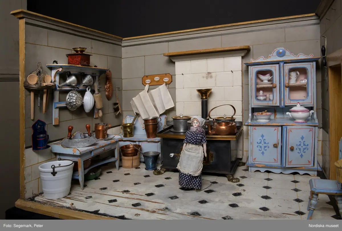 Inventering Sesam 1996-1999:
L 88  H 43,5  Dj 44   cm
Dockskåp, köksdockskåp, av trä, skafferi och kök, utvändigt träådrat. Väggarna i köket kvadermålade och golvet svart och vitrutigt. Skafferiets väggar rosa, väggfasta hyllor, dörrposten ekimiterad.
Lös inredning se inv. 192.992-193.058
Birgitta Martinius 1996