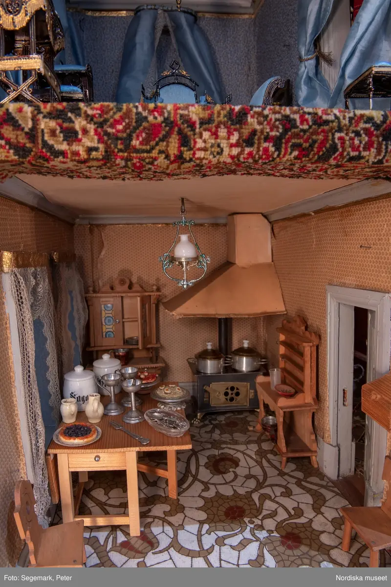 Inventering Sesam 1996-1999:
L 79 cm, B 46 cm, H 174 cm
Dockskåp av brunbetsat furu. Skåp med ett par glasdörrar med brun dekorrand på ramarna, undertill en låda med två dragknoppar, rakt krön med profilerad list. Skåpet står på fyra kulfötter. Dörrparet med lås och nyckel. På gavlarna 4 st fönster på vardera sidan. I skåpet 4 st våningar med sammanlagt 7 rum. Samtliga rum med mönstrade papperstapeter och krok i taket för lampa.
VÅNING 1: 1 rum, heltäckningsmatta av blåmönstrat tyg på vit botten. På höger kortvägg 3 st små pappersbilder i färgtryckav liten flicka i olika situationer. På  bakväggen en europakarta med tysk text.
VÅNING 2:  2 st rum. Bägge med mönstrade linoleummattor och två fönster. Gråmålad dörr mellan rummen. Vänster rum; Spiskåpa av vit papp på bakväggen. 2 st gardinlufter av vit spets med kornischer av gulmetall med påklistrad guldpappersremsa med mönster i relief. Höger rum; två gardinlufter av brunrandigt tyg, kornischer som ovan, omtag av metallkedjor på ena luften och rosa sidenband på andra. Ovanför dörren sitter 3 st beslag av pressad plåt. En större med måne och blommor och två mindre med sköld och blommor. 
VÅNING 3: bägge rummen med mönstrad heltäckningsmatta med lugg och två fönster. Vänster rum; 2 gardinlufter, 1 sänghimmel och 1 draperi av blått siden med vit kantspets. Gardiner med kornischer som ovan. 5 st omtag i form av snodd av gulmetall. Höger rum; enfärgade röda tapeter,2 st gardinlufter av rödrandigt siden med omtag av samma tyg, undergardin av vit spets, kornischer som ovan. Ett draperi av rött och randigt (i flera färger) ylle med ett omtag av metallkedja.
VÅNING 4: Vänster rum; flerfärgat och småmönstrad linoleummatta, Höger rum; utan golvbeläggning. 
- Ett par gröna gardiner av stärkt bomull, som ursprunglingen har suttit innanför glasdörrarna på dockskåpet. Rynkade i båda kortändorna. B 46,5, L 128 cm
Har tillhört givaren. (född 1886 enl. adelskalendern). 
Inventarier till dockskåpet inv.nr 164571-164728
Leif Wallin nov 1997