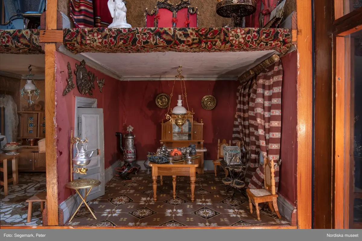 Inventering Sesam 1996-1999:
L 79 cm, B 46 cm, H 174 cm
Dockskåp av brunbetsat furu. Skåp med ett par glasdörrar med brun dekorrand på ramarna, undertill en låda med två dragknoppar, rakt krön med profilerad list. Skåpet står på fyra kulfötter. Dörrparet med lås och nyckel. På gavlarna 4 st fönster på vardera sidan. I skåpet 4 st våningar med sammanlagt 7 rum. Samtliga rum med mönstrade papperstapeter och krok i taket för lampa.
VÅNING 1: 1 rum, heltäckningsmatta av blåmönstrat tyg på vit botten. På höger kortvägg 3 st små pappersbilder i färgtryckav liten flicka i olika situationer. På  bakväggen en europakarta med tysk text.
VÅNING 2:  2 st rum. Bägge med mönstrade linoleummattor och två fönster. Gråmålad dörr mellan rummen. Vänster rum; Spiskåpa av vit papp på bakväggen. 2 st gardinlufter av vit spets med kornischer av gulmetall med påklistrad guldpappersremsa med mönster i relief. Höger rum; två gardinlufter av brunrandigt tyg, kornischer som ovan, omtag av metallkedjor på ena luften och rosa sidenband på andra. Ovanför dörren sitter 3 st beslag av pressad plåt. En större med måne och blommor och två mindre med sköld och blommor. 
VÅNING 3: bägge rummen med mönstrad heltäckningsmatta med lugg och två fönster. Vänster rum; 2 gardinlufter, 1 sänghimmel och 1 draperi av blått siden med vit kantspets. Gardiner med kornischer som ovan. 5 st omtag i form av snodd av gulmetall. Höger rum; enfärgade röda tapeter,2 st gardinlufter av rödrandigt siden med omtag av samma tyg, undergardin av vit spets, kornischer som ovan. Ett draperi av rött och randigt (i flera färger) ylle med ett omtag av metallkedja.
VÅNING 4: Vänster rum; flerfärgat och småmönstrad linoleummatta, Höger rum; utan golvbeläggning. 
- Ett par gröna gardiner av stärkt bomull, som ursprunglingen har suttit innanför glasdörrarna på dockskåpet. Rynkade i båda kortändorna. B 46,5, L 128 cm
Har tillhört givaren. (född 1886 enl. adelskalendern). 
Inventarier till dockskåpet inv.nr 164571-164728
Leif Wallin nov 1997