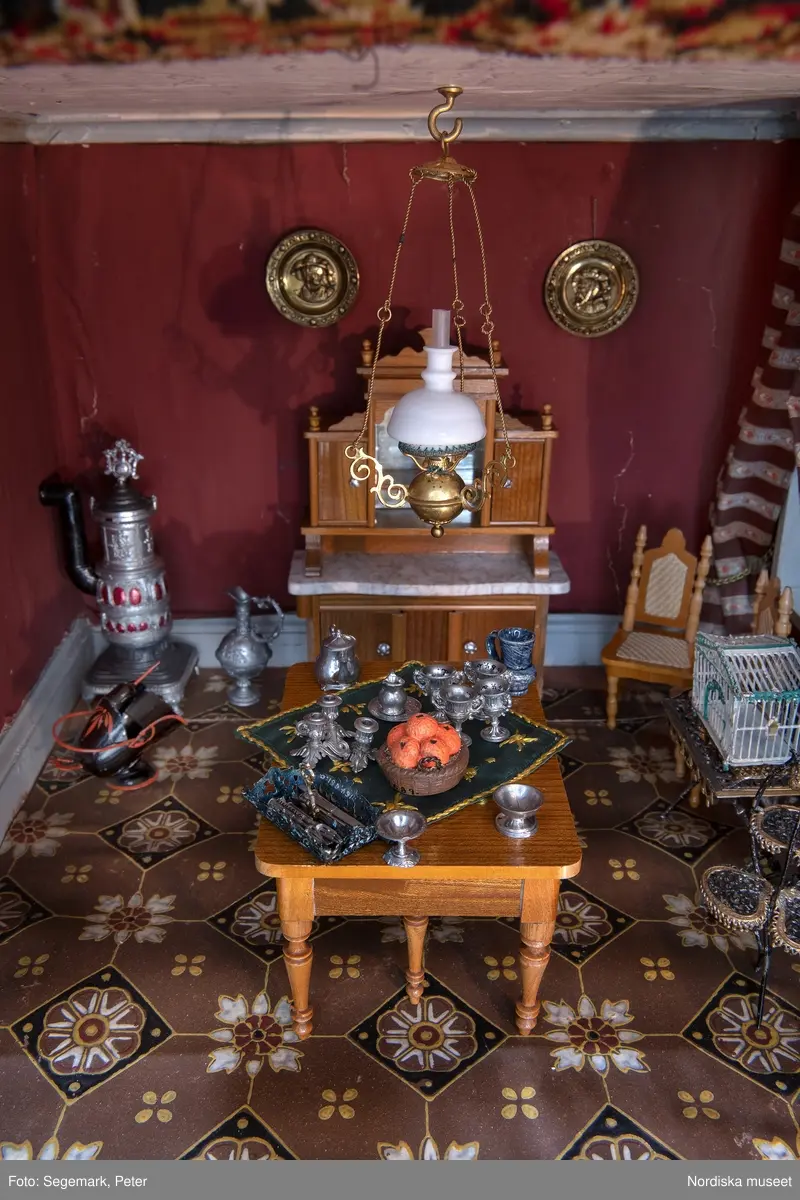 Inventering Sesam 1996-1999:
L 79 cm, B 46 cm, H 174 cm
Dockskåp av brunbetsat furu. Skåp med ett par glasdörrar med brun dekorrand på ramarna, undertill en låda med två dragknoppar, rakt krön med profilerad list. Skåpet står på fyra kulfötter. Dörrparet med lås och nyckel. På gavlarna 4 st fönster på vardera sidan. I skåpet 4 st våningar med sammanlagt 7 rum. Samtliga rum med mönstrade papperstapeter och krok i taket för lampa.
VÅNING 1: 1 rum, heltäckningsmatta av blåmönstrat tyg på vit botten. På höger kortvägg 3 st små pappersbilder i färgtryckav liten flicka i olika situationer. På  bakväggen en europakarta med tysk text.
VÅNING 2:  2 st rum. Bägge med mönstrade linoleummattor och två fönster. Gråmålad dörr mellan rummen. Vänster rum; Spiskåpa av vit papp på bakväggen. 2 st gardinlufter av vit spets med kornischer av gulmetall med påklistrad guldpappersremsa med mönster i relief. Höger rum; två gardinlufter av brunrandigt tyg, kornischer som ovan, omtag av metallkedjor på ena luften och rosa sidenband på andra. Ovanför dörren sitter 3 st beslag av pressad plåt. En större med måne och blommor och två mindre med sköld och blommor. 
VÅNING 3: bägge rummen med mönstrad heltäckningsmatta med lugg och två fönster. Vänster rum; 2 gardinlufter, 1 sänghimmel och 1 draperi av blått siden med vit kantspets. Gardiner med kornischer som ovan. 5 st omtag i form av snodd av gulmetall. Höger rum; enfärgade röda tapeter,2 st gardinlufter av rödrandigt siden med omtag av samma tyg, undergardin av vit spets, kornischer som ovan. Ett draperi av rött och randigt (i flera färger) ylle med ett omtag av metallkedja.
VÅNING 4: Vänster rum; flerfärgat och småmönstrad linoleummatta, Höger rum; utan golvbeläggning. 
- Ett par gröna gardiner av stärkt bomull, som ursprunglingen har suttit innanför glasdörrarna på dockskåpet. Rynkade i båda kortändorna. B 46,5, L 128 cm
Har tillhört givaren. (född 1886 enl. adelskalendern). 
Inventarier till dockskåpet inv.nr 164571-164728
Leif Wallin nov 1997