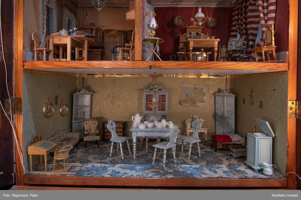 Inventering Sesam 1996-1999:
L 79 cm, B 46 cm, H 174 cm
Dockskåp av brunbetsat furu. Skåp med ett par glasdörrar med brun dekorrand på ramarna, undertill en låda med två dragknoppar, rakt krön med profilerad list. Skåpet står på fyra kulfötter. Dörrparet med lås och nyckel. På gavlarna 4 st fönster på vardera sidan. I skåpet 4 st våningar med sammanlagt 7 rum. Samtliga rum med mönstrade papperstapeter och krok i taket för lampa.
VÅNING 1: 1 rum, heltäckningsmatta av blåmönstrat tyg på vit botten. På höger kortvägg 3 st små pappersbilder i färgtryckav liten flicka i olika situationer. På  bakväggen en europakarta med tysk text.
VÅNING 2:  2 st rum. Bägge med mönstrade linoleummattor och två fönster. Gråmålad dörr mellan rummen. Vänster rum; Spiskåpa av vit papp på bakväggen. 2 st gardinlufter av vit spets med kornischer av gulmetall med påklistrad guldpappersremsa med mönster i relief. Höger rum; två gardinlufter av brunrandigt tyg, kornischer som ovan, omtag av metallkedjor på ena luften och rosa sidenband på andra. Ovanför dörren sitter 3 st beslag av pressad plåt. En större med måne och blommor och två mindre med sköld och blommor. 
VÅNING 3: bägge rummen med mönstrad heltäckningsmatta med lugg och två fönster. Vänster rum; 2 gardinlufter, 1 sänghimmel och 1 draperi av blått siden med vit kantspets. Gardiner med kornischer som ovan. 5 st omtag i form av snodd av gulmetall. Höger rum; enfärgade röda tapeter,2 st gardinlufter av rödrandigt siden med omtag av samma tyg, undergardin av vit spets, kornischer som ovan. Ett draperi av rött och randigt (i flera färger) ylle med ett omtag av metallkedja.
VÅNING 4: Vänster rum; flerfärgat och småmönstrad linoleummatta, Höger rum; utan golvbeläggning. 
- Ett par gröna gardiner av stärkt bomull, som ursprunglingen har suttit innanför glasdörrarna på dockskåpet. Rynkade i båda kortändorna. B 46,5, L 128 cm
Har tillhört givaren. (född 1886 enl. adelskalendern). 
Inventarier till dockskåpet inv.nr 164571-164728
Leif Wallin nov 1997