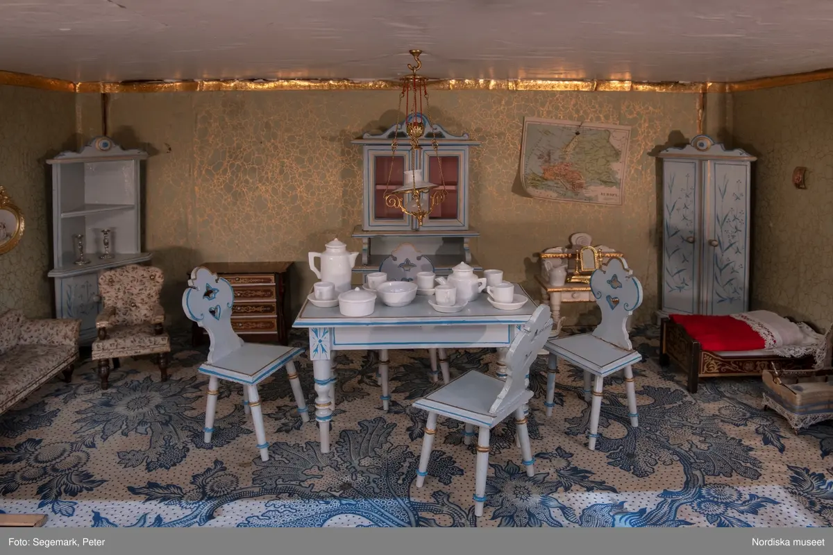 Inventering Sesam 1996-1999:
L 79 cm, B 46 cm, H 174 cm
Dockskåp av brunbetsat furu. Skåp med ett par glasdörrar med brun dekorrand på ramarna, undertill en låda med två dragknoppar, rakt krön med profilerad list. Skåpet står på fyra kulfötter. Dörrparet med lås och nyckel. På gavlarna 4 st fönster på vardera sidan. I skåpet 4 st våningar med sammanlagt 7 rum. Samtliga rum med mönstrade papperstapeter och krok i taket för lampa.
VÅNING 1: 1 rum, heltäckningsmatta av blåmönstrat tyg på vit botten. På höger kortvägg 3 st små pappersbilder i färgtryckav liten flicka i olika situationer. På  bakväggen en europakarta med tysk text.
VÅNING 2:  2 st rum. Bägge med mönstrade linoleummattor och två fönster. Gråmålad dörr mellan rummen. Vänster rum; Spiskåpa av vit papp på bakväggen. 2 st gardinlufter av vit spets med kornischer av gulmetall med påklistrad guldpappersremsa med mönster i relief. Höger rum; två gardinlufter av brunrandigt tyg, kornischer som ovan, omtag av metallkedjor på ena luften och rosa sidenband på andra. Ovanför dörren sitter 3 st beslag av pressad plåt. En större med måne och blommor och två mindre med sköld och blommor. 
VÅNING 3: bägge rummen med mönstrad heltäckningsmatta med lugg och två fönster. Vänster rum; 2 gardinlufter, 1 sänghimmel och 1 draperi av blått siden med vit kantspets. Gardiner med kornischer som ovan. 5 st omtag i form av snodd av gulmetall. Höger rum; enfärgade röda tapeter,2 st gardinlufter av rödrandigt siden med omtag av samma tyg, undergardin av vit spets, kornischer som ovan. Ett draperi av rött och randigt (i flera färger) ylle med ett omtag av metallkedja.
VÅNING 4: Vänster rum; flerfärgat och småmönstrad linoleummatta, Höger rum; utan golvbeläggning. 
- Ett par gröna gardiner av stärkt bomull, som ursprunglingen har suttit innanför glasdörrarna på dockskåpet. Rynkade i båda kortändorna. B 46,5, L 128 cm
Har tillhört givaren. (född 1886 enl. adelskalendern). 
Inventarier till dockskåpet inv.nr 164571-164728
Leif Wallin nov 1997