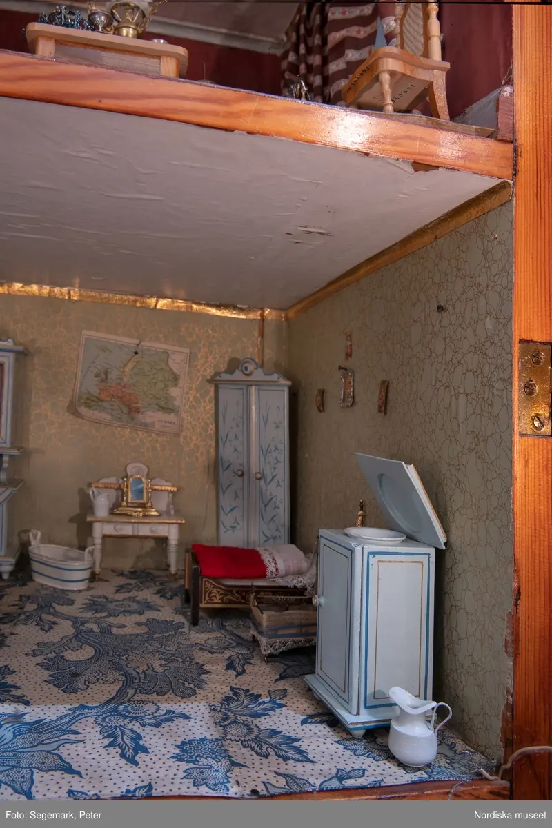 Inventering Sesam 1996-1999:
L 79 cm, B 46 cm, H 174 cm
Dockskåp av brunbetsat furu. Skåp med ett par glasdörrar med brun dekorrand på ramarna, undertill en låda med två dragknoppar, rakt krön med profilerad list. Skåpet står på fyra kulfötter. Dörrparet med lås och nyckel. På gavlarna 4 st fönster på vardera sidan. I skåpet 4 st våningar med sammanlagt 7 rum. Samtliga rum med mönstrade papperstapeter och krok i taket för lampa.
VÅNING 1: 1 rum, heltäckningsmatta av blåmönstrat tyg på vit botten. På höger kortvägg 3 st små pappersbilder i färgtryckav liten flicka i olika situationer. På  bakväggen en europakarta med tysk text.
VÅNING 2:  2 st rum. Bägge med mönstrade linoleummattor och två fönster. Gråmålad dörr mellan rummen. Vänster rum; Spiskåpa av vit papp på bakväggen. 2 st gardinlufter av vit spets med kornischer av gulmetall med påklistrad guldpappersremsa med mönster i relief. Höger rum; två gardinlufter av brunrandigt tyg, kornischer som ovan, omtag av metallkedjor på ena luften och rosa sidenband på andra. Ovanför dörren sitter 3 st beslag av pressad plåt. En större med måne och blommor och två mindre med sköld och blommor. 
VÅNING 3: bägge rummen med mönstrad heltäckningsmatta med lugg och två fönster. Vänster rum; 2 gardinlufter, 1 sänghimmel och 1 draperi av blått siden med vit kantspets. Gardiner med kornischer som ovan. 5 st omtag i form av snodd av gulmetall. Höger rum; enfärgade röda tapeter,2 st gardinlufter av rödrandigt siden med omtag av samma tyg, undergardin av vit spets, kornischer som ovan. Ett draperi av rött och randigt (i flera färger) ylle med ett omtag av metallkedja.
VÅNING 4: Vänster rum; flerfärgat och småmönstrad linoleummatta, Höger rum; utan golvbeläggning. 
- Ett par gröna gardiner av stärkt bomull, som ursprunglingen har suttit innanför glasdörrarna på dockskåpet. Rynkade i båda kortändorna. B 46,5, L 128 cm
Har tillhört givaren. (född 1886 enl. adelskalendern). 
Inventarier till dockskåpet inv.nr 164571-164728
Leif Wallin nov 1997