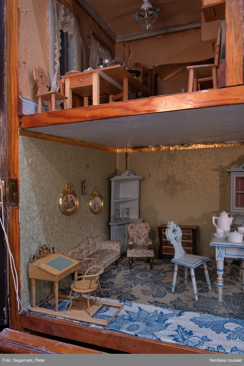 Inventering Sesam 1996-1999:
L 79 cm, B 46 cm, H 174 cm
Dockskåp av brunbetsat furu. Skåp med ett par glasdörrar med brun dekorrand på ramarna, undertill en låda med två dragknoppar, rakt krön med profilerad list. Skåpet står på fyra kulfötter. Dörrparet med lås och nyckel. På gavlarna 4 st fönster på vardera sidan. I skåpet 4 st våningar med sammanlagt 7 rum. Samtliga rum med mönstrade papperstapeter och krok i taket för lampa.
VÅNING 1: 1 rum, heltäckningsmatta av blåmönstrat tyg på vit botten. På höger kortvägg 3 st små pappersbilder i färgtryckav liten flicka i olika situationer. På  bakväggen en europakarta med tysk text.
VÅNING 2:  2 st rum. Bägge med mönstrade linoleummattor och två fönster. Gråmålad dörr mellan rummen. Vänster rum; Spiskåpa av vit papp på bakväggen. 2 st gardinlufter av vit spets med kornischer av gulmetall med påklistrad guldpappersremsa med mönster i relief. Höger rum; två gardinlufter av brunrandigt tyg, kornischer som ovan, omtag av metallkedjor på ena luften och rosa sidenband på andra. Ovanför dörren sitter 3 st beslag av pressad plåt. En större med måne och blommor och två mindre med sköld och blommor. 
VÅNING 3: bägge rummen med mönstrad heltäckningsmatta med lugg och två fönster. Vänster rum; 2 gardinlufter, 1 sänghimmel och 1 draperi av blått siden med vit kantspets. Gardiner med kornischer som ovan. 5 st omtag i form av snodd av gulmetall. Höger rum; enfärgade röda tapeter,2 st gardinlufter av rödrandigt siden med omtag av samma tyg, undergardin av vit spets, kornischer som ovan. Ett draperi av rött och randigt (i flera färger) ylle med ett omtag av metallkedja.
VÅNING 4: Vänster rum; flerfärgat och småmönstrad linoleummatta, Höger rum; utan golvbeläggning. 
- Ett par gröna gardiner av stärkt bomull, som ursprunglingen har suttit innanför glasdörrarna på dockskåpet. Rynkade i båda kortändorna. B 46,5, L 128 cm
Har tillhört givaren. (född 1886 enl. adelskalendern). 
Inventarier till dockskåpet inv.nr 164571-164728
Leif Wallin nov 1997