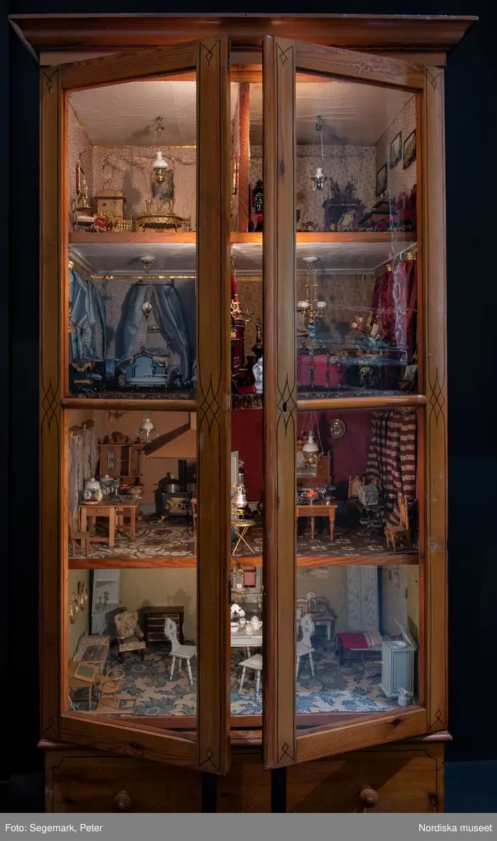 Inventering Sesam 1996-1999:
L 79 cm, B 46 cm, H 174 cm
Dockskåp av brunbetsat furu. Skåp med ett par glasdörrar med brun dekorrand på ramarna, undertill en låda med två dragknoppar, rakt krön med profilerad list. Skåpet står på fyra kulfötter. Dörrparet med lås och nyckel. På gavlarna 4 st fönster på vardera sidan. I skåpet 4 st våningar med sammanlagt 7 rum. Samtliga rum med mönstrade papperstapeter och krok i taket för lampa.
VÅNING 1: 1 rum, heltäckningsmatta av blåmönstrat tyg på vit botten. På höger kortvägg 3 st små pappersbilder i färgtryckav liten flicka i olika situationer. På  bakväggen en europakarta med tysk text.
VÅNING 2:  2 st rum. Bägge med mönstrade linoleummattor och två fönster. Gråmålad dörr mellan rummen. Vänster rum; Spiskåpa av vit papp på bakväggen. 2 st gardinlufter av vit spets med kornischer av gulmetall med påklistrad guldpappersremsa med mönster i relief. Höger rum; två gardinlufter av brunrandigt tyg, kornischer som ovan, omtag av metallkedjor på ena luften och rosa sidenband på andra. Ovanför dörren sitter 3 st beslag av pressad plåt. En större med måne och blommor och två mindre med sköld och blommor. 
VÅNING 3: bägge rummen med mönstrad heltäckningsmatta med lugg och två fönster. Vänster rum; 2 gardinlufter, 1 sänghimmel och 1 draperi av blått siden med vit kantspets. Gardiner med kornischer som ovan. 5 st omtag i form av snodd av gulmetall. Höger rum; enfärgade röda tapeter,2 st gardinlufter av rödrandigt siden med omtag av samma tyg, undergardin av vit spets, kornischer som ovan. Ett draperi av rött och randigt (i flera färger) ylle med ett omtag av metallkedja.
VÅNING 4: Vänster rum; flerfärgat och småmönstrad linoleummatta, Höger rum; utan golvbeläggning. 
- Ett par gröna gardiner av stärkt bomull, som ursprunglingen har suttit innanför glasdörrarna på dockskåpet. Rynkade i båda kortändorna. B 46,5, L 128 cm
Har tillhört givaren. (född 1886 enl. adelskalendern). 
Inventarier till dockskåpet inv.nr 164571-164728
Leif Wallin nov 1997