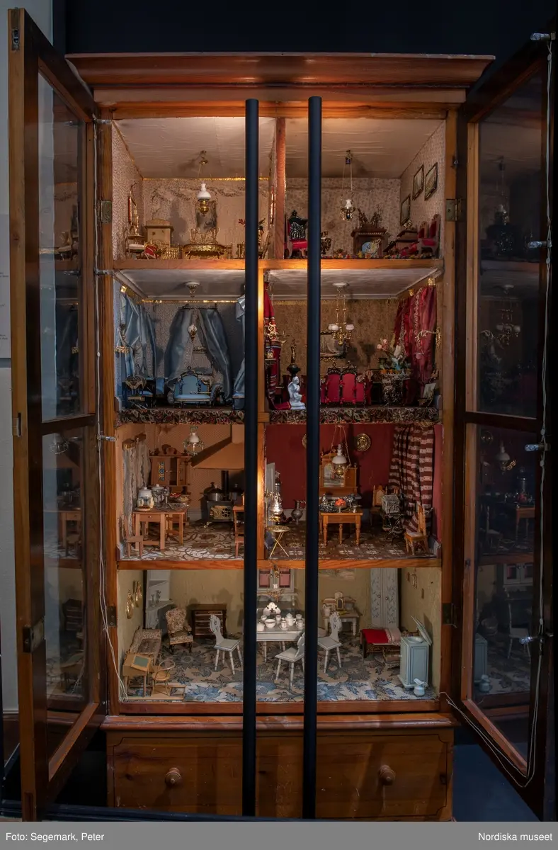 Inventering Sesam 1996-1999:
L 79 cm, B 46 cm, H 174 cm
Dockskåp av brunbetsat furu. Skåp med ett par glasdörrar med brun dekorrand på ramarna, undertill en låda med två dragknoppar, rakt krön med profilerad list. Skåpet står på fyra kulfötter. Dörrparet med lås och nyckel. På gavlarna 4 st fönster på vardera sidan. I skåpet 4 st våningar med sammanlagt 7 rum. Samtliga rum med mönstrade papperstapeter och krok i taket för lampa.
VÅNING 1: 1 rum, heltäckningsmatta av blåmönstrat tyg på vit botten. På höger kortvägg 3 st små pappersbilder i färgtryckav liten flicka i olika situationer. På  bakväggen en europakarta med tysk text.
VÅNING 2:  2 st rum. Bägge med mönstrade linoleummattor och två fönster. Gråmålad dörr mellan rummen. Vänster rum; Spiskåpa av vit papp på bakväggen. 2 st gardinlufter av vit spets med kornischer av gulmetall med påklistrad guldpappersremsa med mönster i relief. Höger rum; två gardinlufter av brunrandigt tyg, kornischer som ovan, omtag av metallkedjor på ena luften och rosa sidenband på andra. Ovanför dörren sitter 3 st beslag av pressad plåt. En större med måne och blommor och två mindre med sköld och blommor. 
VÅNING 3: bägge rummen med mönstrad heltäckningsmatta med lugg och två fönster. Vänster rum; 2 gardinlufter, 1 sänghimmel och 1 draperi av blått siden med vit kantspets. Gardiner med kornischer som ovan. 5 st omtag i form av snodd av gulmetall. Höger rum; enfärgade röda tapeter,2 st gardinlufter av rödrandigt siden med omtag av samma tyg, undergardin av vit spets, kornischer som ovan. Ett draperi av rött och randigt (i flera färger) ylle med ett omtag av metallkedja.
VÅNING 4: Vänster rum; flerfärgat och småmönstrad linoleummatta, Höger rum; utan golvbeläggning. 
- Ett par gröna gardiner av stärkt bomull, som ursprunglingen har suttit innanför glasdörrarna på dockskåpet. Rynkade i båda kortändorna. B 46,5, L 128 cm
Har tillhört givaren. (född 1886 enl. adelskalendern). 
Inventarier till dockskåpet inv.nr 164571-164728
Leif Wallin nov 1997