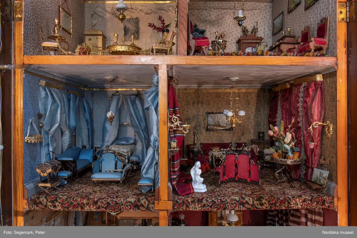 Inventering Sesam 1996-1999:
L 79 cm, B 46 cm, H 174 cm
Dockskåp av brunbetsat furu. Skåp med ett par glasdörrar med brun dekorrand på ramarna, undertill en låda med två dragknoppar, rakt krön med profilerad list. Skåpet står på fyra kulfötter. Dörrparet med lås och nyckel. På gavlarna 4 st fönster på vardera sidan. I skåpet 4 st våningar med sammanlagt 7 rum. Samtliga rum med mönstrade papperstapeter och krok i taket för lampa.
VÅNING 1: 1 rum, heltäckningsmatta av blåmönstrat tyg på vit botten. På höger kortvägg 3 st små pappersbilder i färgtryckav liten flicka i olika situationer. På  bakväggen en europakarta med tysk text.
VÅNING 2:  2 st rum. Bägge med mönstrade linoleummattor och två fönster. Gråmålad dörr mellan rummen. Vänster rum; Spiskåpa av vit papp på bakväggen. 2 st gardinlufter av vit spets med kornischer av gulmetall med påklistrad guldpappersremsa med mönster i relief. Höger rum; två gardinlufter av brunrandigt tyg, kornischer som ovan, omtag av metallkedjor på ena luften och rosa sidenband på andra. Ovanför dörren sitter 3 st beslag av pressad plåt. En större med måne och blommor och två mindre med sköld och blommor. 
VÅNING 3: bägge rummen med mönstrad heltäckningsmatta med lugg och två fönster. Vänster rum; 2 gardinlufter, 1 sänghimmel och 1 draperi av blått siden med vit kantspets. Gardiner med kornischer som ovan. 5 st omtag i form av snodd av gulmetall. Höger rum; enfärgade röda tapeter,2 st gardinlufter av rödrandigt siden med omtag av samma tyg, undergardin av vit spets, kornischer som ovan. Ett draperi av rött och randigt (i flera färger) ylle med ett omtag av metallkedja.
VÅNING 4: Vänster rum; flerfärgat och småmönstrad linoleummatta, Höger rum; utan golvbeläggning. 
- Ett par gröna gardiner av stärkt bomull, som ursprunglingen har suttit innanför glasdörrarna på dockskåpet. Rynkade i båda kortändorna. B 46,5, L 128 cm
Har tillhört givaren. (född 1886 enl. adelskalendern). 
Inventarier till dockskåpet inv.nr 164571-164728
Leif Wallin nov 1997