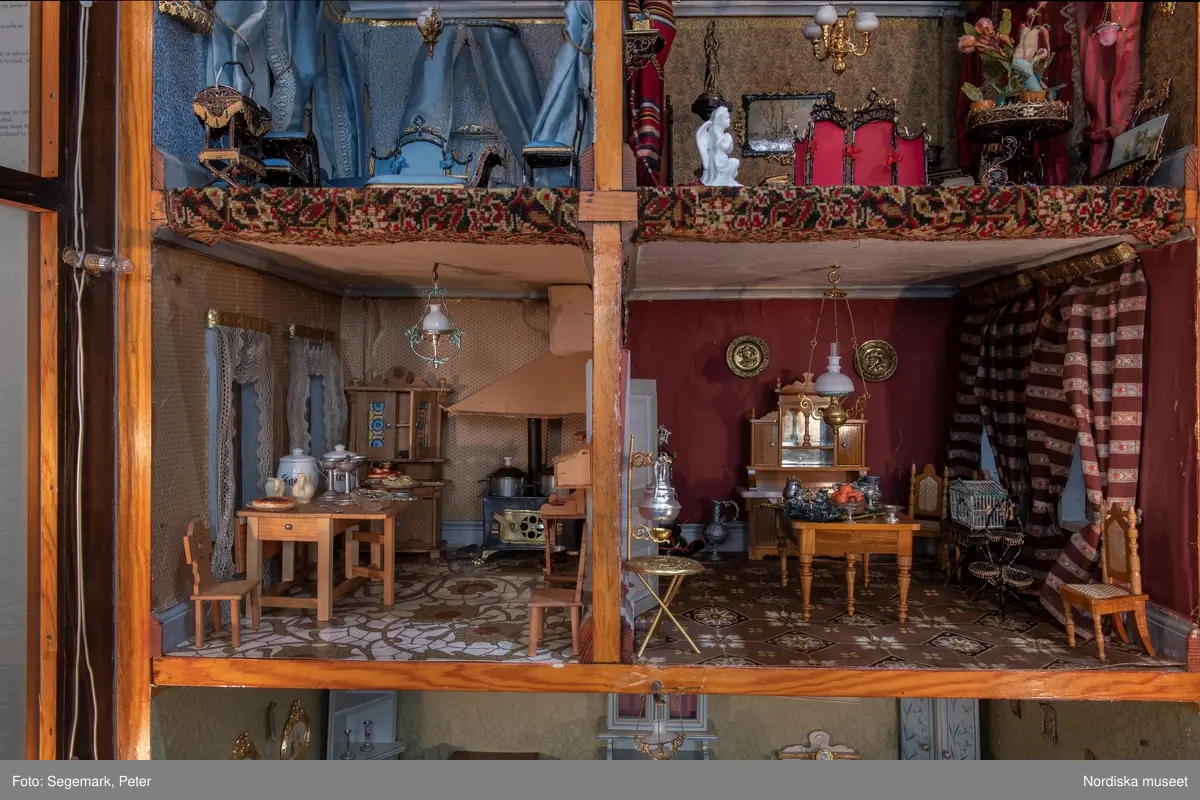 Inventering Sesam 1996-1999:
L 79 cm, B 46 cm, H 174 cm
Dockskåp av brunbetsat furu. Skåp med ett par glasdörrar med brun dekorrand på ramarna, undertill en låda med två dragknoppar, rakt krön med profilerad list. Skåpet står på fyra kulfötter. Dörrparet med lås och nyckel. På gavlarna 4 st fönster på vardera sidan. I skåpet 4 st våningar med sammanlagt 7 rum. Samtliga rum med mönstrade papperstapeter och krok i taket för lampa.
VÅNING 1: 1 rum, heltäckningsmatta av blåmönstrat tyg på vit botten. På höger kortvägg 3 st små pappersbilder i färgtryckav liten flicka i olika situationer. På  bakväggen en europakarta med tysk text.
VÅNING 2:  2 st rum. Bägge med mönstrade linoleummattor och två fönster. Gråmålad dörr mellan rummen. Vänster rum; Spiskåpa av vit papp på bakväggen. 2 st gardinlufter av vit spets med kornischer av gulmetall med påklistrad guldpappersremsa med mönster i relief. Höger rum; två gardinlufter av brunrandigt tyg, kornischer som ovan, omtag av metallkedjor på ena luften och rosa sidenband på andra. Ovanför dörren sitter 3 st beslag av pressad plåt. En större med måne och blommor och två mindre med sköld och blommor. 
VÅNING 3: bägge rummen med mönstrad heltäckningsmatta med lugg och två fönster. Vänster rum; 2 gardinlufter, 1 sänghimmel och 1 draperi av blått siden med vit kantspets. Gardiner med kornischer som ovan. 5 st omtag i form av snodd av gulmetall. Höger rum; enfärgade röda tapeter,2 st gardinlufter av rödrandigt siden med omtag av samma tyg, undergardin av vit spets, kornischer som ovan. Ett draperi av rött och randigt (i flera färger) ylle med ett omtag av metallkedja.
VÅNING 4: Vänster rum; flerfärgat och småmönstrad linoleummatta, Höger rum; utan golvbeläggning. 
- Ett par gröna gardiner av stärkt bomull, som ursprunglingen har suttit innanför glasdörrarna på dockskåpet. Rynkade i båda kortändorna. B 46,5, L 128 cm
Har tillhört givaren. (född 1886 enl. adelskalendern). 
Inventarier till dockskåpet inv.nr 164571-164728
Leif Wallin nov 1997