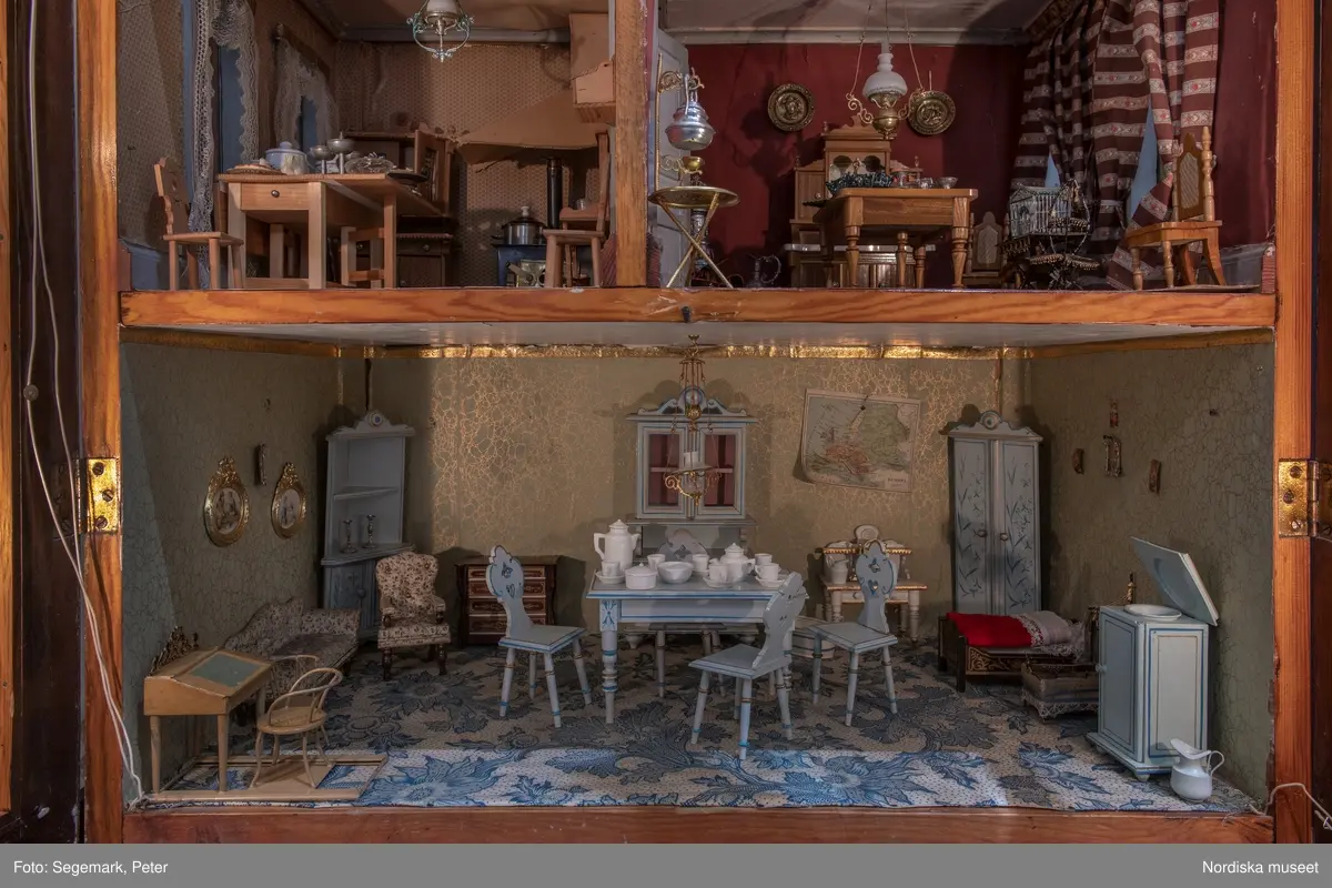 Inventering Sesam 1996-1999:
L 79 cm, B 46 cm, H 174 cm
Dockskåp av brunbetsat furu. Skåp med ett par glasdörrar med brun dekorrand på ramarna, undertill en låda med två dragknoppar, rakt krön med profilerad list. Skåpet står på fyra kulfötter. Dörrparet med lås och nyckel. På gavlarna 4 st fönster på vardera sidan. I skåpet 4 st våningar med sammanlagt 7 rum. Samtliga rum med mönstrade papperstapeter och krok i taket för lampa.
VÅNING 1: 1 rum, heltäckningsmatta av blåmönstrat tyg på vit botten. På höger kortvägg 3 st små pappersbilder i färgtryckav liten flicka i olika situationer. På  bakväggen en europakarta med tysk text.
VÅNING 2:  2 st rum. Bägge med mönstrade linoleummattor och två fönster. Gråmålad dörr mellan rummen. Vänster rum; Spiskåpa av vit papp på bakväggen. 2 st gardinlufter av vit spets med kornischer av gulmetall med påklistrad guldpappersremsa med mönster i relief. Höger rum; två gardinlufter av brunrandigt tyg, kornischer som ovan, omtag av metallkedjor på ena luften och rosa sidenband på andra. Ovanför dörren sitter 3 st beslag av pressad plåt. En större med måne och blommor och två mindre med sköld och blommor. 
VÅNING 3: bägge rummen med mönstrad heltäckningsmatta med lugg och två fönster. Vänster rum; 2 gardinlufter, 1 sänghimmel och 1 draperi av blått siden med vit kantspets. Gardiner med kornischer som ovan. 5 st omtag i form av snodd av gulmetall. Höger rum; enfärgade röda tapeter,2 st gardinlufter av rödrandigt siden med omtag av samma tyg, undergardin av vit spets, kornischer som ovan. Ett draperi av rött och randigt (i flera färger) ylle med ett omtag av metallkedja.
VÅNING 4: Vänster rum; flerfärgat och småmönstrad linoleummatta, Höger rum; utan golvbeläggning. 
- Ett par gröna gardiner av stärkt bomull, som ursprunglingen har suttit innanför glasdörrarna på dockskåpet. Rynkade i båda kortändorna. B 46,5, L 128 cm
Har tillhört givaren. (född 1886 enl. adelskalendern). 
Inventarier till dockskåpet inv.nr 164571-164728
Leif Wallin nov 1997