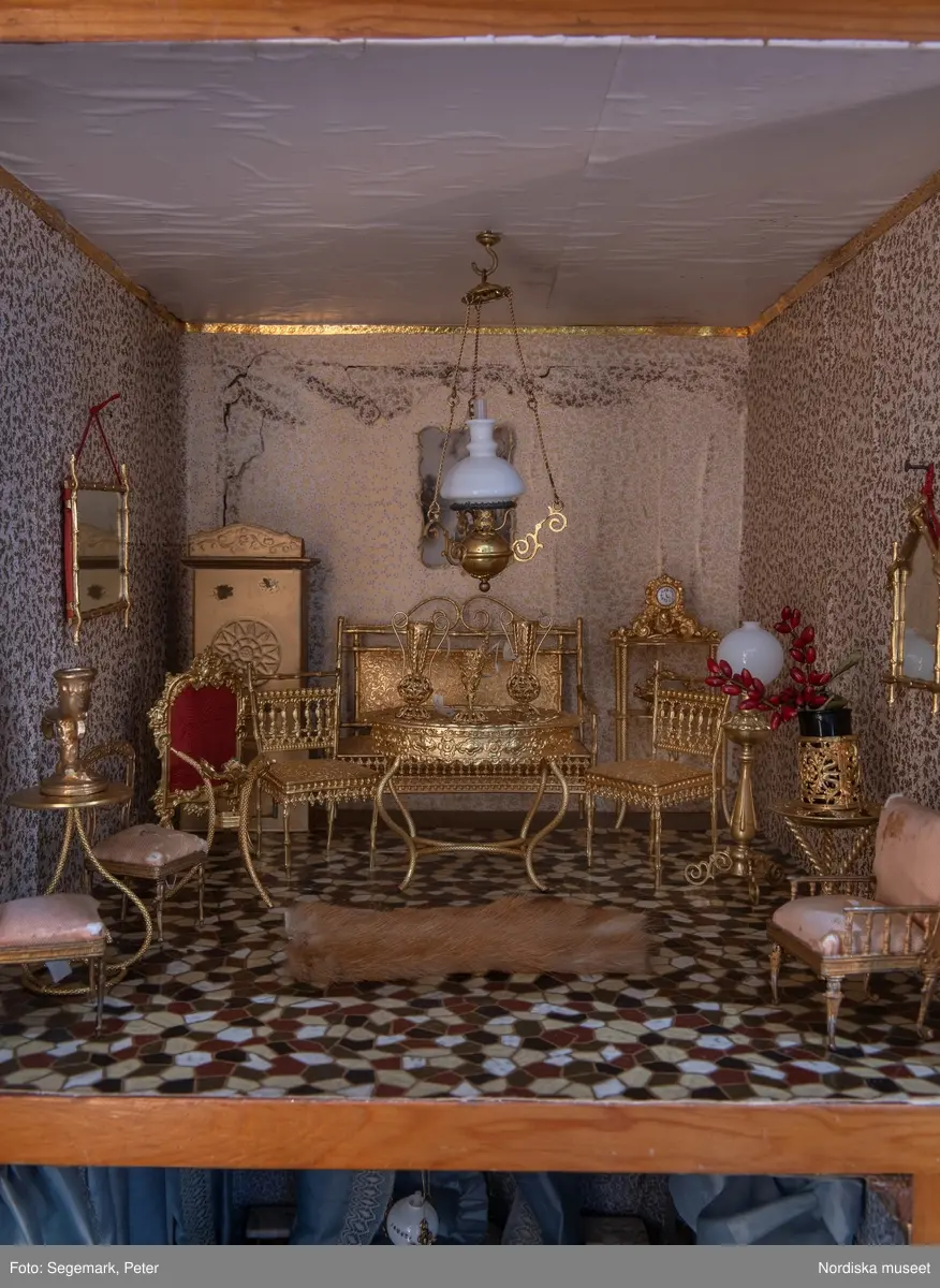 Inventering Sesam 1996-1999:
L 79 cm, B 46 cm, H 174 cm
Dockskåp av brunbetsat furu. Skåp med ett par glasdörrar med brun dekorrand på ramarna, undertill en låda med två dragknoppar, rakt krön med profilerad list. Skåpet står på fyra kulfötter. Dörrparet med lås och nyckel. På gavlarna 4 st fönster på vardera sidan. I skåpet 4 st våningar med sammanlagt 7 rum. Samtliga rum med mönstrade papperstapeter och krok i taket för lampa.
VÅNING 1: 1 rum, heltäckningsmatta av blåmönstrat tyg på vit botten. På höger kortvägg 3 st små pappersbilder i färgtryckav liten flicka i olika situationer. På  bakväggen en europakarta med tysk text.
VÅNING 2:  2 st rum. Bägge med mönstrade linoleummattor och två fönster. Gråmålad dörr mellan rummen. Vänster rum; Spiskåpa av vit papp på bakväggen. 2 st gardinlufter av vit spets med kornischer av gulmetall med påklistrad guldpappersremsa med mönster i relief. Höger rum; två gardinlufter av brunrandigt tyg, kornischer som ovan, omtag av metallkedjor på ena luften och rosa sidenband på andra. Ovanför dörren sitter 3 st beslag av pressad plåt. En större med måne och blommor och två mindre med sköld och blommor. 
VÅNING 3: bägge rummen med mönstrad heltäckningsmatta med lugg och två fönster. Vänster rum; 2 gardinlufter, 1 sänghimmel och 1 draperi av blått siden med vit kantspets. Gardiner med kornischer som ovan. 5 st omtag i form av snodd av gulmetall. Höger rum; enfärgade röda tapeter,2 st gardinlufter av rödrandigt siden med omtag av samma tyg, undergardin av vit spets, kornischer som ovan. Ett draperi av rött och randigt (i flera färger) ylle med ett omtag av metallkedja.
VÅNING 4: Vänster rum; flerfärgat och småmönstrad linoleummatta, Höger rum; utan golvbeläggning. 
- Ett par gröna gardiner av stärkt bomull, som ursprunglingen har suttit innanför glasdörrarna på dockskåpet. Rynkade i båda kortändorna. B 46,5, L 128 cm
Har tillhört givaren. (född 1886 enl. adelskalendern). 
Inventarier till dockskåpet inv.nr 164571-164728
Leif Wallin nov 1997