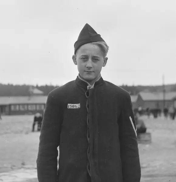 Fotografi tatt ved frigjøringen, i svartt/hvitt. En ung gutt står midt i bildets fokus, vendt mot fotografen. Han har fangedrakt og fangelue på. På fangedrakten står fangenummeret hans: 19062. I bakgrunnen kan man skimte leiren, med flere brakker og fanger.