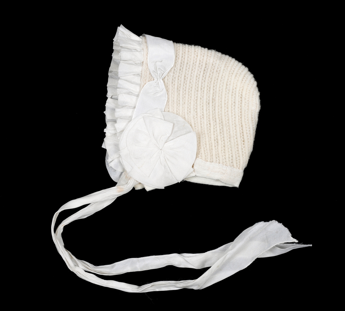 Strikket lue med skru av foldet silke, silkebånd og rosetter over ørene. Fôret med gauze. Kantet i nedkant med gauze. Knytebånd i silke.