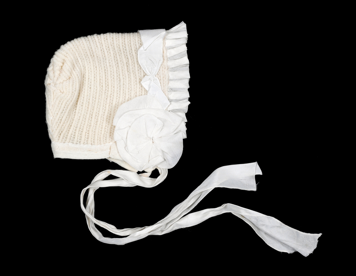 Strikket lue med skru av foldet silke, silkebånd og rosetter over ørene. Fôret med gauze. Kantet i nedkant med gauze. Knytebånd i silke.