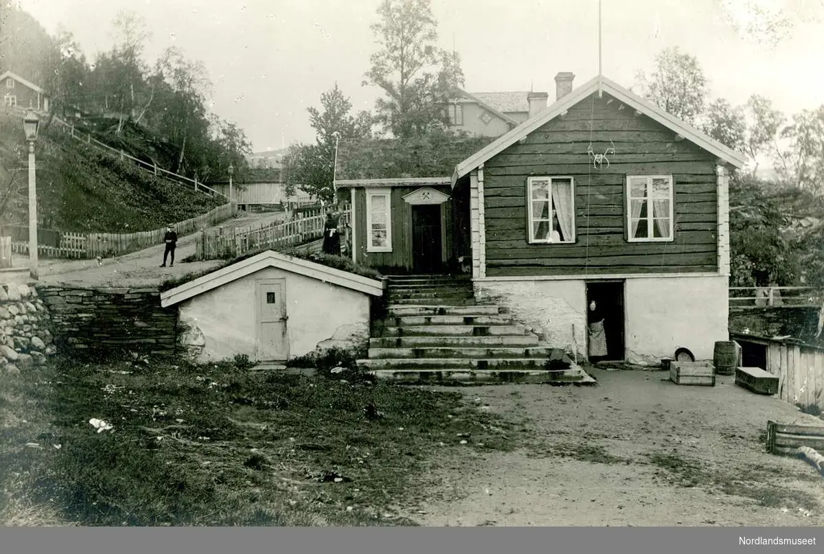 Furulund. 
Som bilde 0482. 

Da Hasselbom kom til Langvandsdalen med sine menn, i et forrykende uvær, den 27. januar 1887 var det ikke mange boliger å søke seg husvære i. Vi vet om et bygg nede ved vannet som Vattenbygdens kobberverk hadde bygd knappe ti år tidligere, ellers var det bare de forskjellige rydningsbrukene rundt om. Men de sto han av som det heter, men hvordan vet vi ikke. Muligens bodde de i telt, de gjorde det den gang også. (Den gang da jernbanen opp til Fossen ble bygget (1891-92) er det beretninger om at det var flere som da bodde langs banen i telt, også gjennom vinteren.)  Men da det tok til å våres i bakkene så ble det tid til byggearbeid også og vi vet at noen av de ansatte bygde seg sine egne hjem. Hasselbom satte i gang bygging av et litt større hus sommeren 1887. 

Det ble omtalt som Messa og det huset ble bygget med en kjelleretasje. I kjelleren ble det innredet et kjøkken og et spiserom. Over kjelleren ble det laftet et hus med en etasje og et loft, eller ”trev” som de sa. I første etasje var det to rom, ett for Hasselbom og et for noen av arbeiderne. De som ikke hadde plass i første etasje holdt til oppe på trevet. I det rommet som Hasselbom hadde var det foruten til hans opphold også kontor, lager og en liten handel. (Det var langt ned til Venset og handelsmannen.) Messa som ble bygget av marerial fra nærområdet ble ferdig i 1888. 

Vi ser her Messa fra ca 1892. Vi ser at den ble laftet av ca. 4” saget plank og bygningen har fått et vindfang og har også en låst matbod ved siden av. Det ser ut til at det har blitt orden omkring bygningen, til å med veilys langs veien. Bak messa ser vi boligen til direktøren, bak der igjen er det en stall og oppe i bakken et hus som inneholdt bolig, lager og et arrestlokale. (Og friområdene, ser vi er inngjerdet med stakittgjerde mot folk eller dyr?) 

Foto Ukjent.