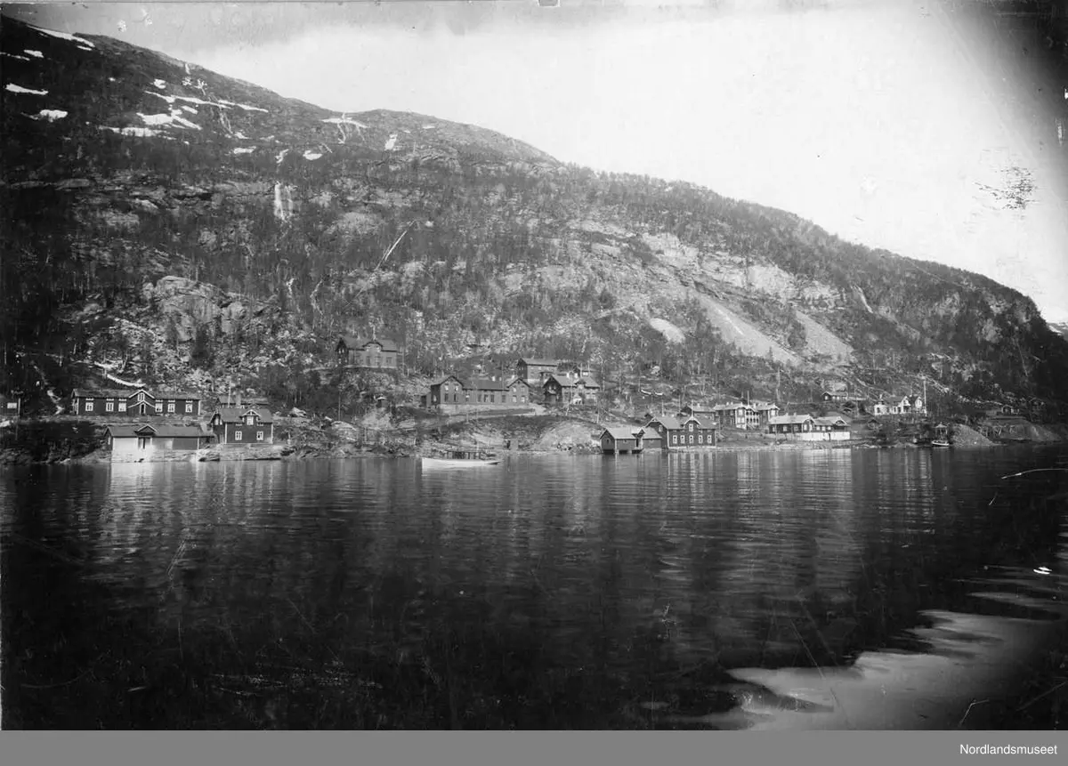 Furulund. 
Den sentrale delen sett fra vannet. Foran ligger ”Gutten” stille ute på vannet. 

Fra venstre ses Mellageret og Bakeriet. Bakom er Brakke 3 og Brakke 2. (Det kan virke som at brakke 2 i en periode ble benyttet som bakeri med et tilbygg som mellager ved siden av inntil nytt bakeri og mellager senere ble bygget nede ved vannet i 1898? 

Den første bakeren som er kjent ansatt ved bedriften, etter at gruveselskapet var begynt å vokse til noe stort ca 1890, var Bakermester Sievert Rise fra Oppdal. Han hadde tidligere drevet eget bakeri i Viktoriahavn. Ettersom arbeidet på jernbanen der nå var stanset så ville han få seg arbeid et annet sted. Med hans søknad fulgte både attest og svarporto.) 

Så er det Sykehuset ovenfor Dampkjøkkenet og Direksjonsvillaen med ”Villa Mons Petter” ovenfor den sist nevnte. Nede ved vannet er Butikken på kaia med den sjøboden som var varelageret til butikken. Over taket til butikken ses øvre del av den gamle butikken vest for kontoret som nå var ombygget til ingeniørbolig. 

Så kommer Messa og Direktørboligen med en lagerbyggning til venstre oppe i bakken, bygget av stein. Det huset rommet i tillegg til et lager også en familiebolig og et arrestlokale. Helt til høyre er det et lite hus, høyst sannsynlig er det Voll-brakka som senere fikk navnet Mjåseth-brakka. Lengre bak kan det skimtes en bygning og da er vi oppe på Verkstedhaugen. 

Tidspunktet kan være 1900/1901. 

Foto Ukjent.