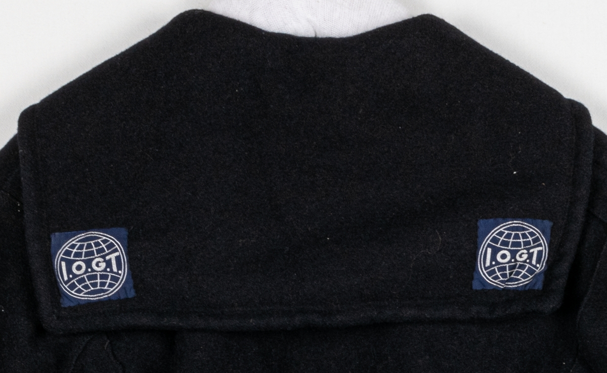 Djupt mörkblå bussarong med sjömanskrage. Två fickor på bröstet samt tygemblem med svensk flagga. Kragen har två fastsydda tygemblem med texten "I.O.G.T.".
