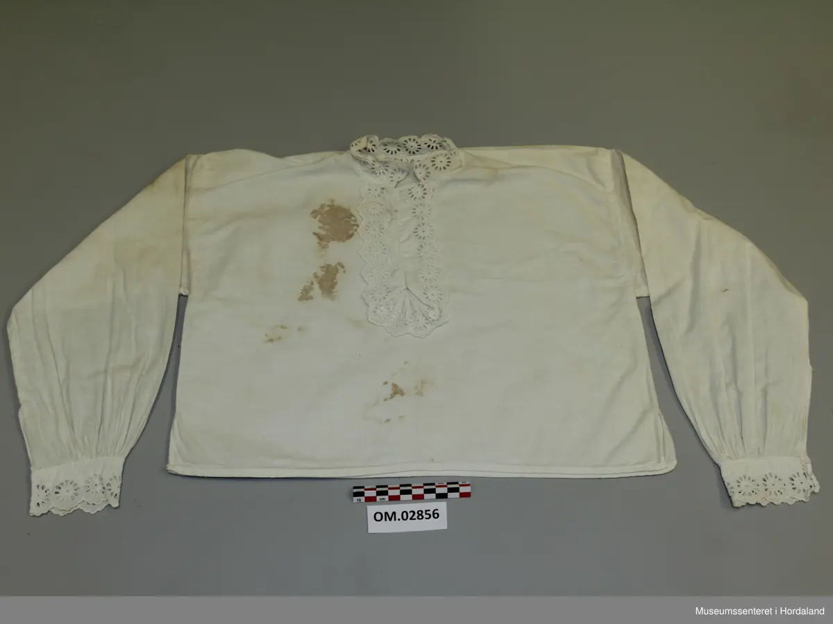 Kvit skjorte med blondekantar til jente. Rett modell med rynking ved armmansjettane. Blonder i bringa med knapping under. Store brune flekkar.