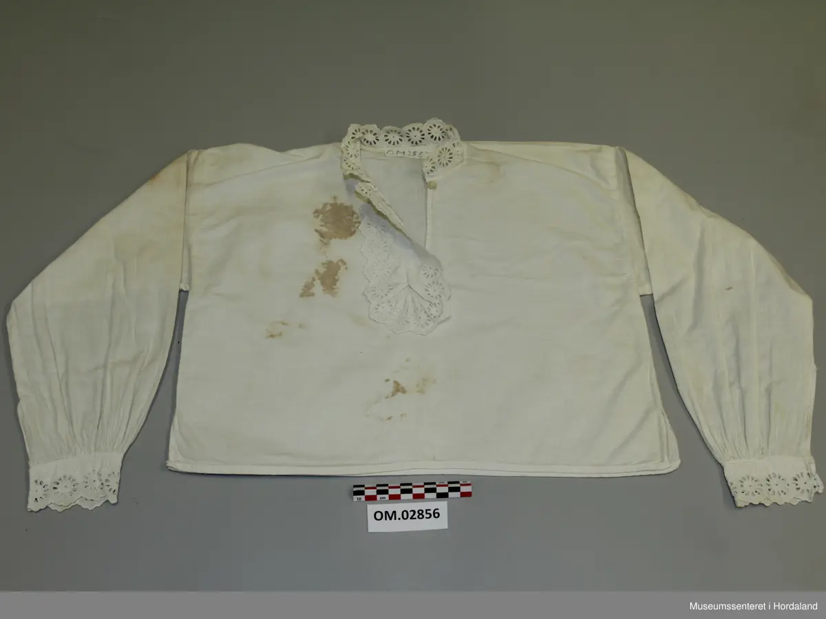 Kvit skjorte med blondekantar til jente. Rett modell med rynking ved armmansjettane. Blonder i bringa med knapping under. Store brune flekkar.