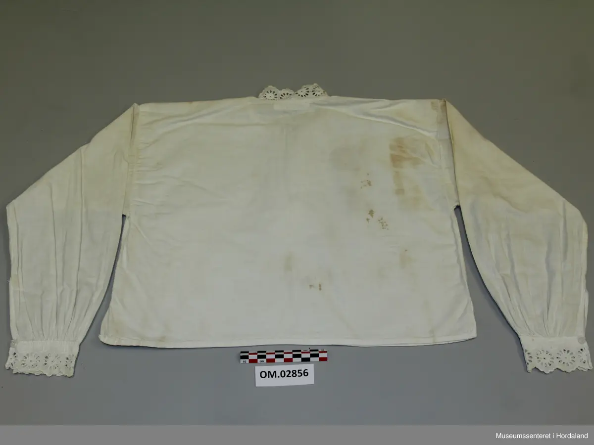 Kvit skjorte med blondekantar til jente. Rett modell med rynking ved armmansjettane. Blonder i bringa med knapping under. Store brune flekkar.