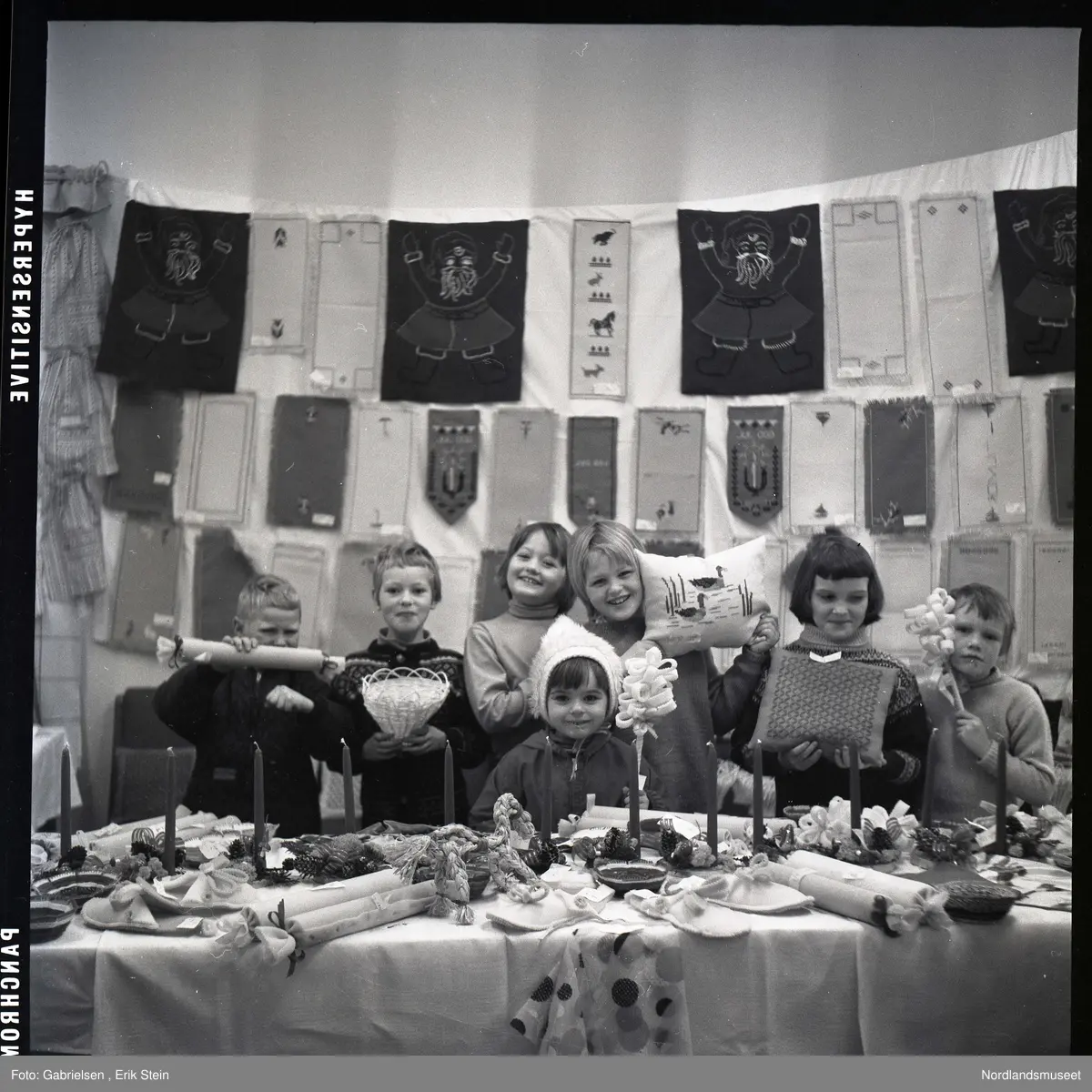 Fotografi av en gruppe med barn som selger puter med mønster og julelys og kurver og kjevler og juleduker og noen tøfler og andre ting på et langt bord i et rom med et vindu med noen gardiner et ukjent sted