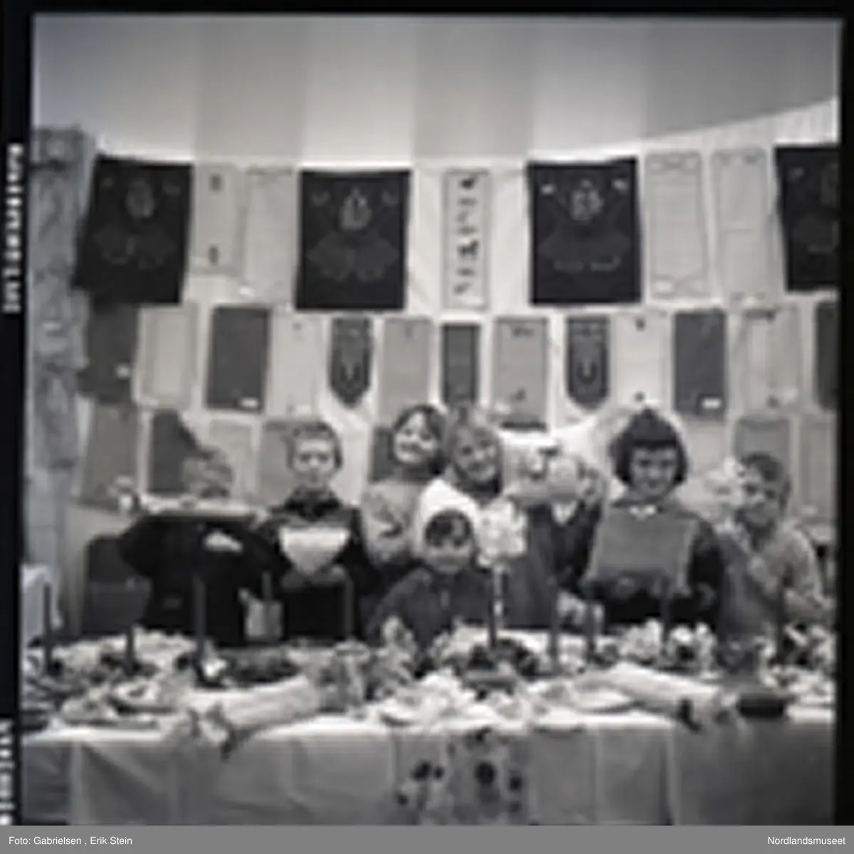 Fotografi av en gruppe med barn som selger puter med mønster og julelys og kurver og kjevler og juleduker og noen tøfler og andre ting på et langt bord i et rom med et vindu med noen gardiner et ukjent sted