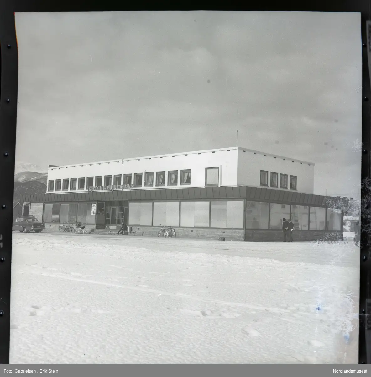 Fotografi av Rognan samvirkelag med mange vinduer 
og en hovedinngang med to dører og et skilt 
over innganget på taket som det står samvirkelaget
på og noen biler som står parkert utenfor samvirkelaget 
og en snøslette vedsiden av parkeringsplassen til samvirkelaget
der man ser det går to menn kledd i vinterklær og hatter 
og man ser en mann som sitter på trappen til samvirkelaget
og røyker og en li med skoger lenger borte fra Rognan
og et stort fjell og man ser en kvinne som holder en veske
som går fra samvirkelaget over parkeringsplassen mot sentrum
lengst til høyre på bildet
