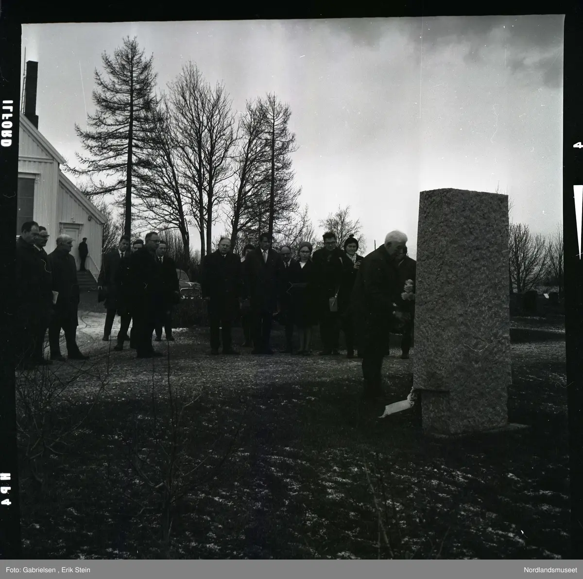 Fotografi av mange finkledde menn og kvinner som står på kirkegården til Fauske kirke og ser på en mann kledd i dress med slips som legger en krans ned på en minnestein 
til falne soldater under andre verdenskrig i Fauske
i en minnesmarkering man ser at de står en mann på trappen
til en dør inn til Fauskekirken og man ser en bil som står parkert vedsiden av Fauskekirken på plenen med gravsteienene
på kirkegården og man ser at det vokser noen trær
rundt i kirkegården