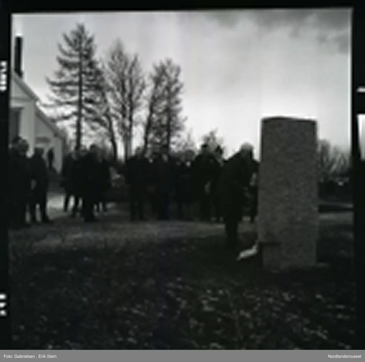 Fotografi av mange finkledde menn og kvinner som står på kirkegården til Fauske kirke og ser på en mann kledd i dress med slips som legger en krans ned på en minnestein 
til falne soldater under andre verdenskrig i Fauske
i en minnesmarkering man ser at de står en mann på trappen
til en dør inn til Fauskekirken og man ser en bil som står parkert vedsiden av Fauskekirken på plenen med gravsteienene
på kirkegården og man ser at det vokser noen trær
rundt i kirkegården