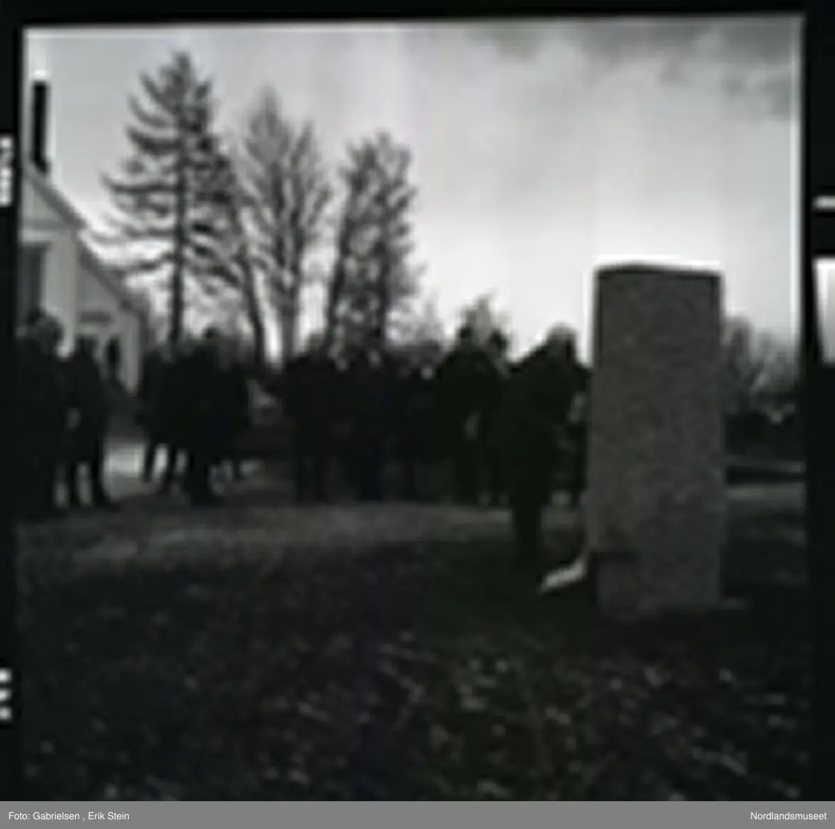 Fotografi av mange finkledde menn og kvinner som står på kirkegården til Fauske kirke og ser på en mann kledd i dress med slips som legger en krans ned på en minnestein 
til falne soldater under andre verdenskrig i Fauske
i en minnesmarkering man ser at de står en mann på trappen
til en dør inn til Fauskekirken og man ser en bil som står parkert vedsiden av Fauskekirken på plenen med gravsteienene
på kirkegården og man ser at det vokser noen trær
rundt i kirkegården