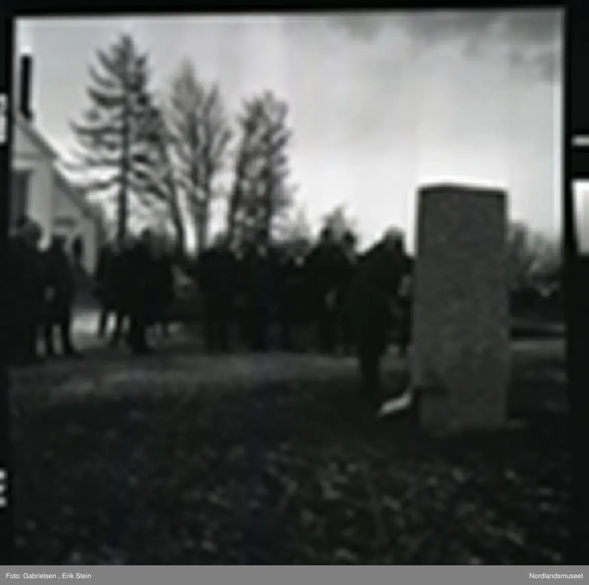 Fotografi av mange finkledde menn og kvinner som står på kirkegården til Fauske kirke og ser på en mann kledd i dress med slips som legger en krans ned på en minnestein 
til falne soldater under andre verdenskrig i Fauske
i en minnesmarkering man ser at de står en mann på trappen
til en dør inn til Fauskekirken og man ser en bil som står parkert vedsiden av Fauskekirken på plenen med gravsteienene
på kirkegården og man ser at det vokser noen trær
rundt i kirkegården
