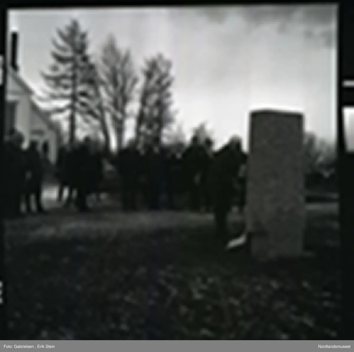 Fotografi av mange finkledde menn og kvinner som står på kirkegården til Fauske kirke og ser på en mann kledd i dress med slips som legger en krans ned på en minnestein 
til falne soldater under andre verdenskrig i Fauske
i en minnesmarkering man ser at de står en mann på trappen
til en dør inn til Fauskekirken og man ser en bil som står parkert vedsiden av Fauskekirken på plenen med gravsteienene
på kirkegården og man ser at det vokser noen trær
rundt i kirkegården