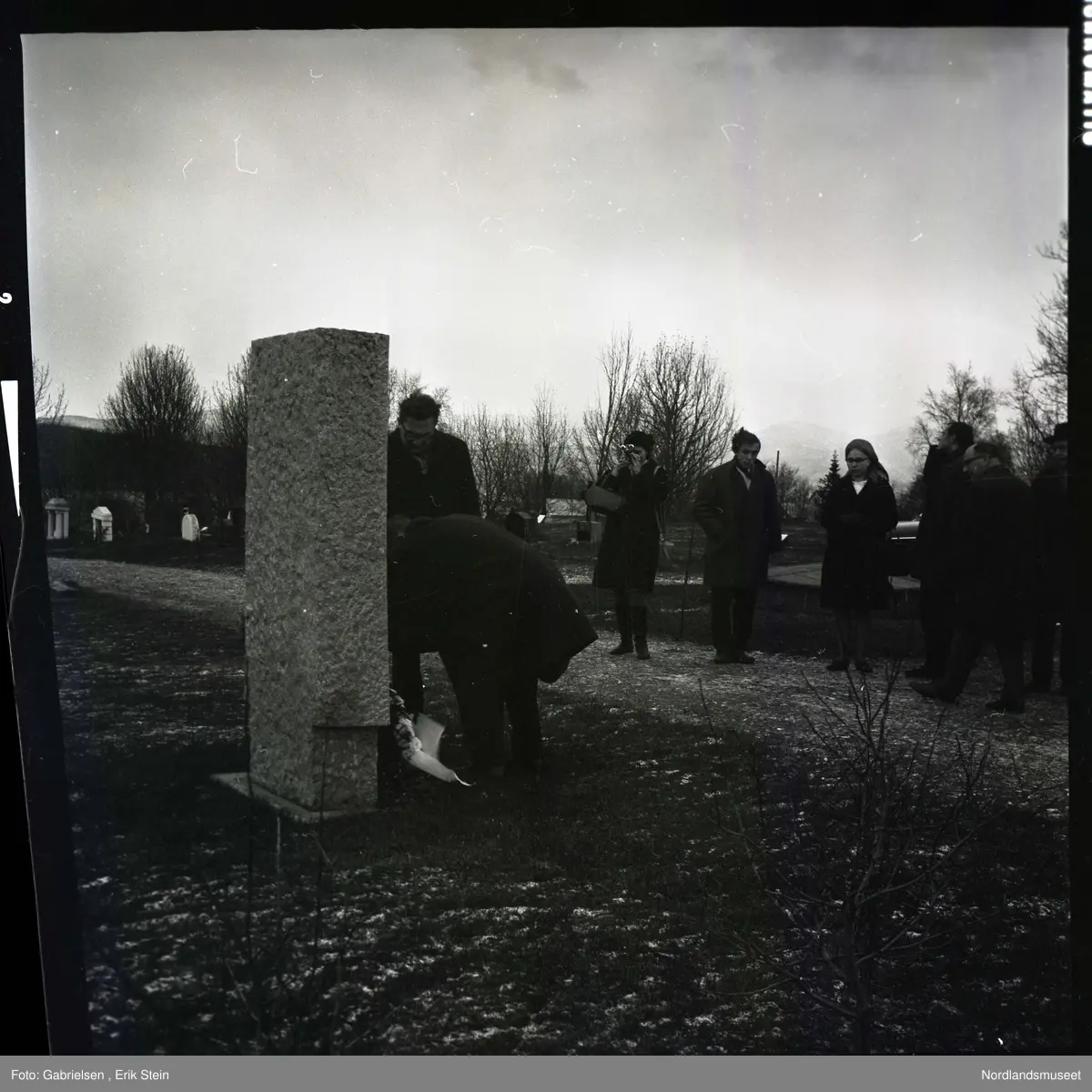 Fotografi av en minnemarkering på Fauske kirkegård 
som det står noen finkledde kvinner og menn og ser på to menn som legger ned en krans på en minnestein 
til falne soldater i Fauske under andre verdenskrig 
og en kvinne som tar bilde av mennen som legger ned kransen 
og man ser en fontene bak mennen og kvinnen i kirkegården
som det ligger et lokk over og man ser noen trær som vokser i kirkegården og en grusvei som går opp til kirkegården 
og mange gravsteiner i kirkegården