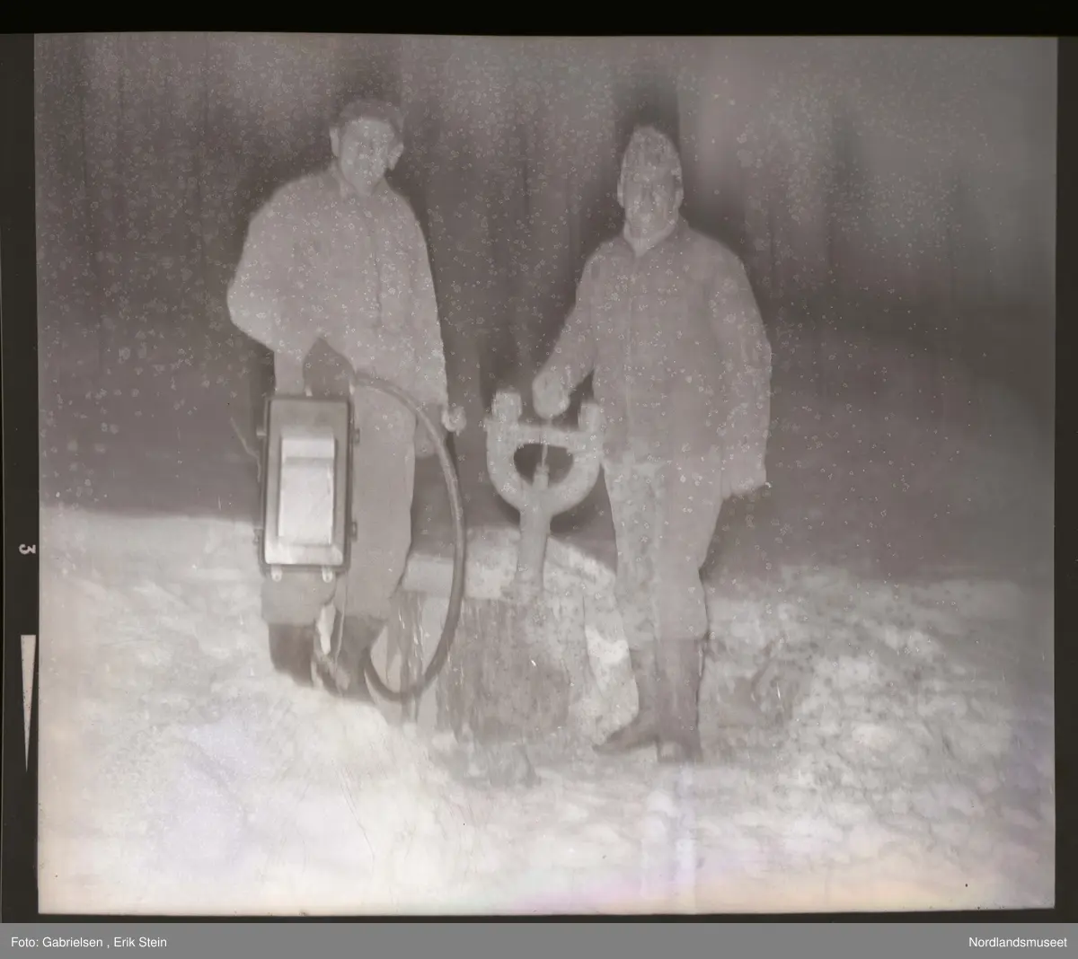 Fotografi av to menn som står i snøen vedsiden av noen rør
til kloakken som de har arbeidet med 
man ser at mannen som står til venstre holder i et håndtak
til en liten maskin med et rør som går til kloakken 
og mannen til høyrer holder i to rør som går ned mot kloakken 
under bakken med mye snø på 
og man ser en skog med noen trær lenger borte i en snøbakke 
og at de er kledd i støvler og luer og votter
og andre vinterklær
